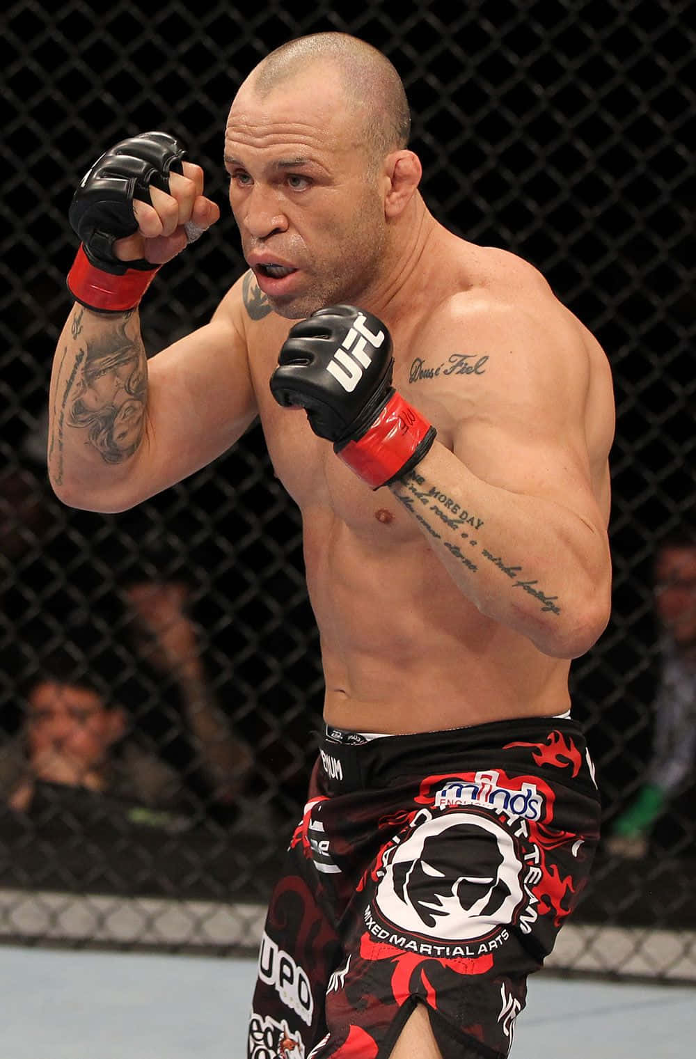 UFC 桌布 壁紙 — 一位肌肉發達的MMA拳手，戴著紅黑手套和短褲，站在籠式八角籠內，手臂上的紋身清晰可見。