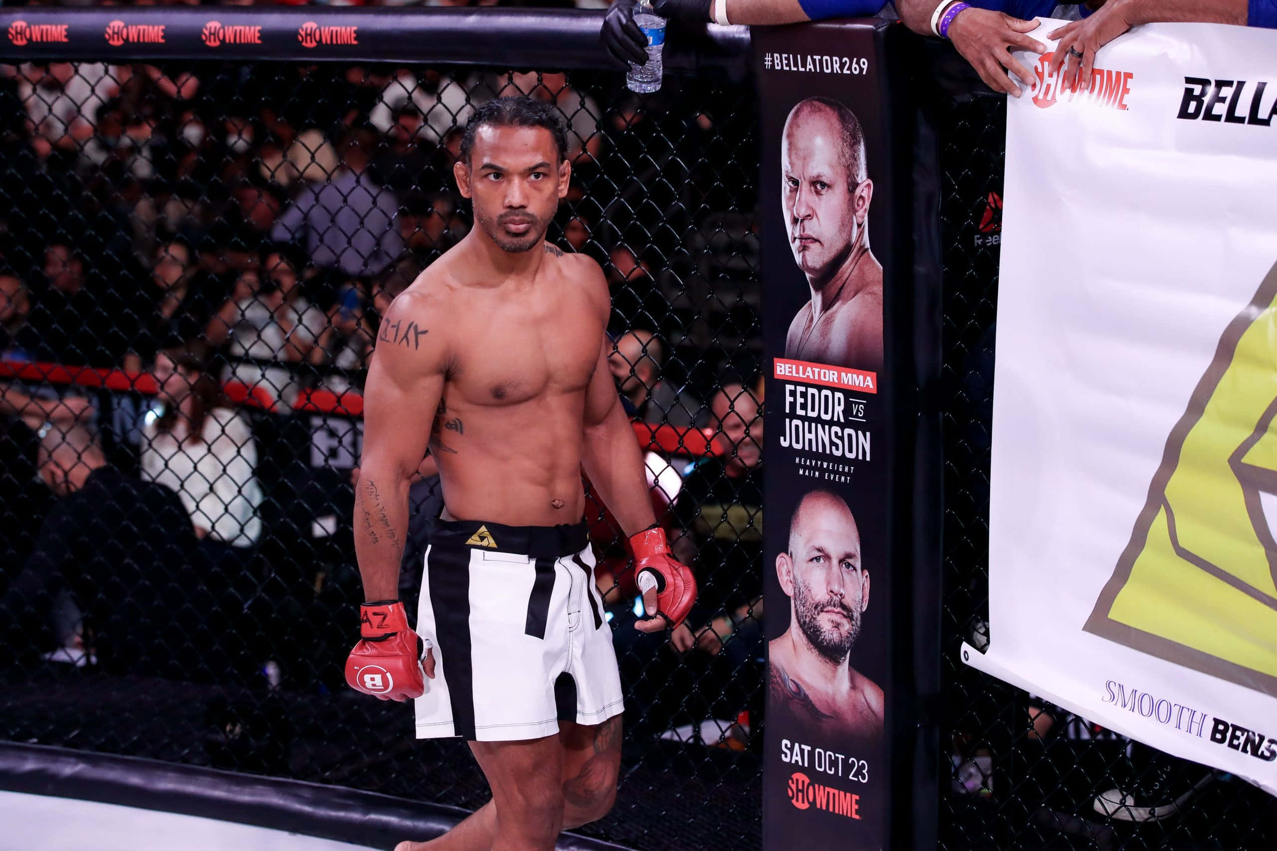 Fight 桌布 壁紙 — 一位肌肉發達的MMA拳手，戴著紅色手套，穿著黑白短褲，站在八角籠內，圍欄後面有群眾。