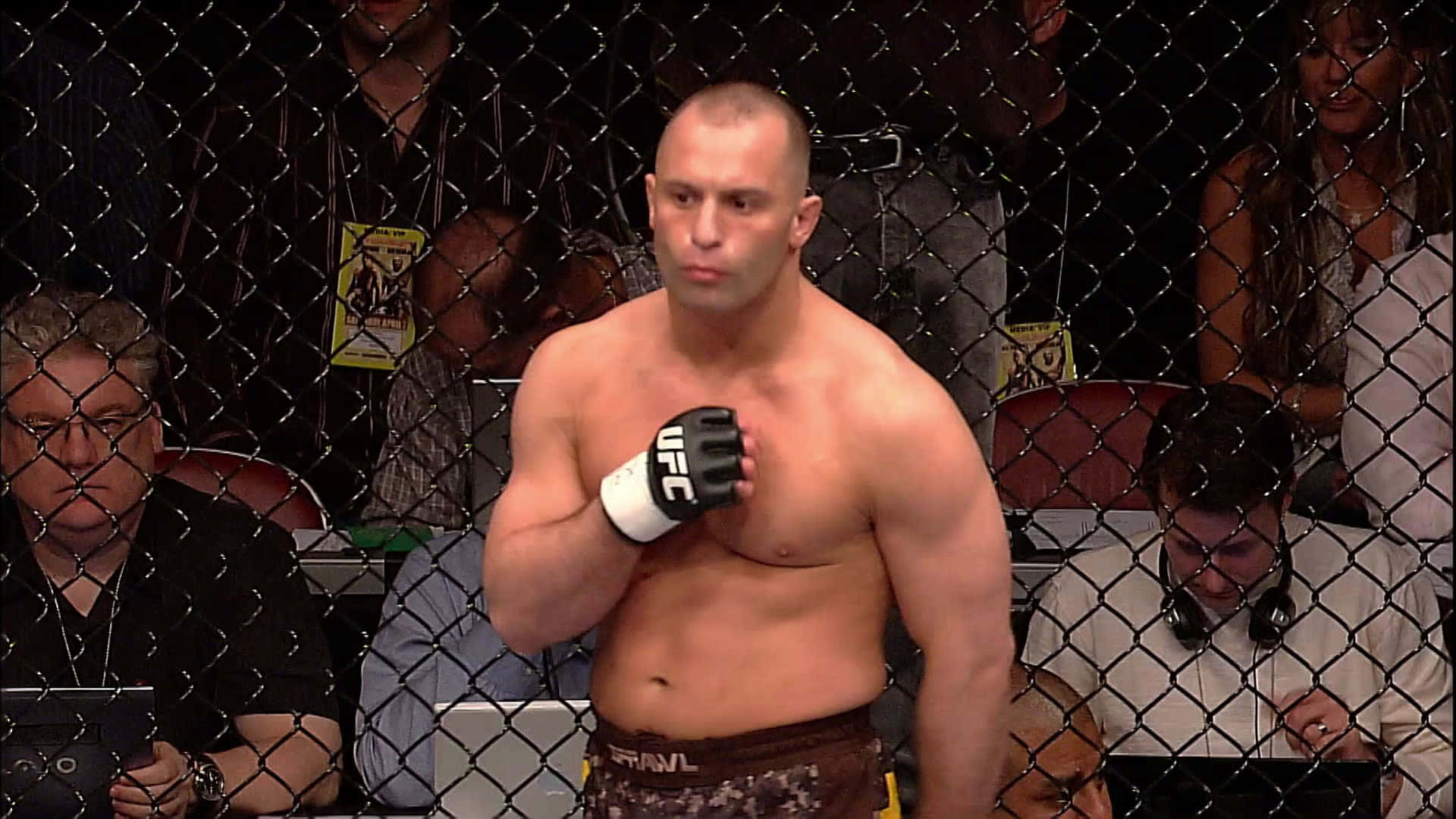 UFC 桌布 壁紙 — 一位肌肉發達的MMA拳手戴著UFC手套，站在八角籠內，觀眾專注地觀看。