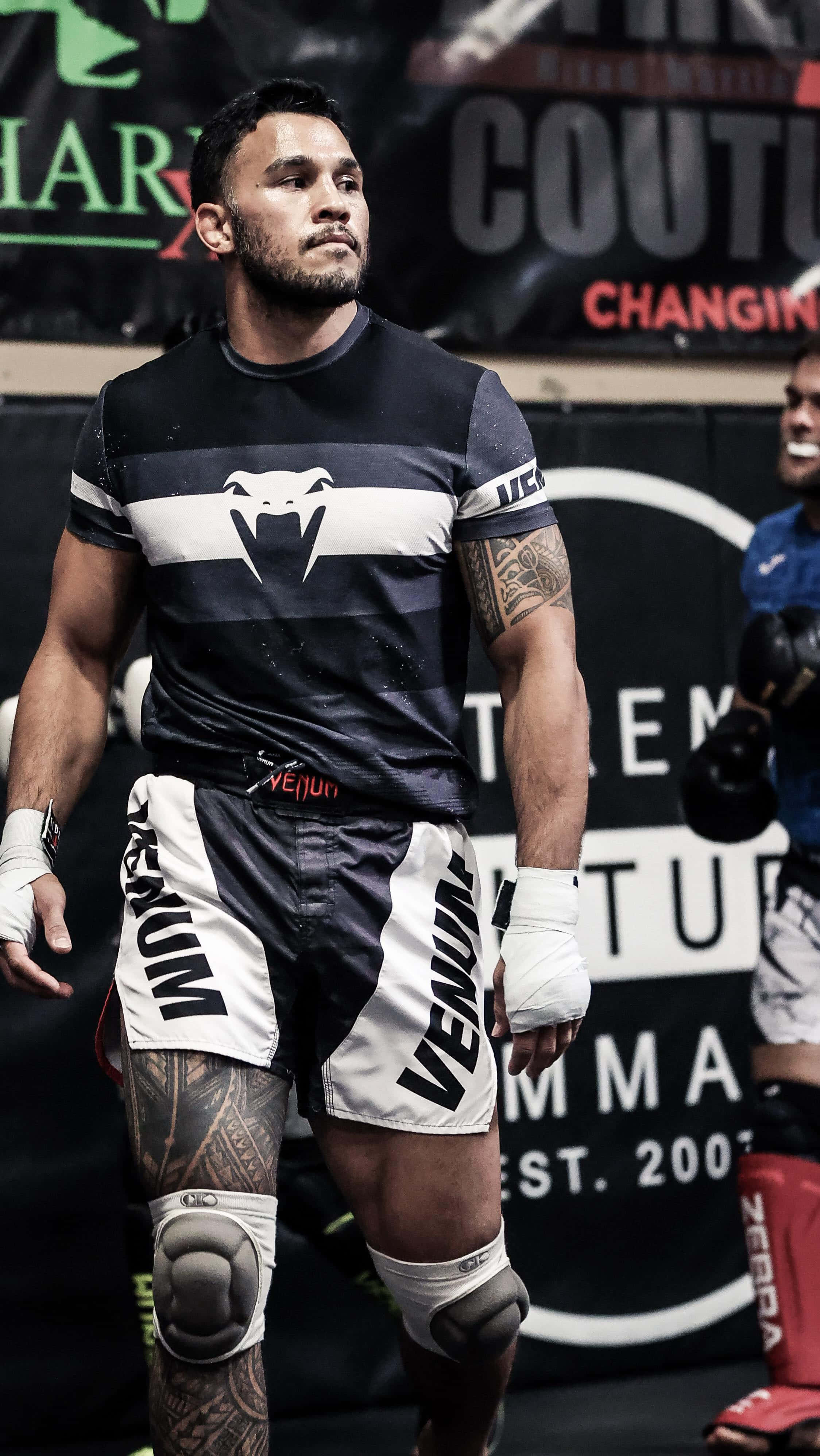 UFC 桌布 壁紙 — 一位肌肉發達的MMA拳手穿著黑白Venum裝備，專注地站在健身房，紋身和護膝清晰可見。