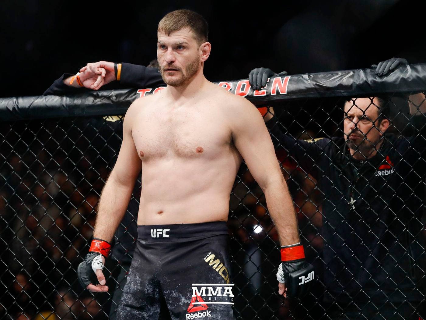 UFC 桌布 壁紙 — 一位肌肉發達的MMA拳手站在籠內，穿著黑色UFC短褲和手套，神情專注。