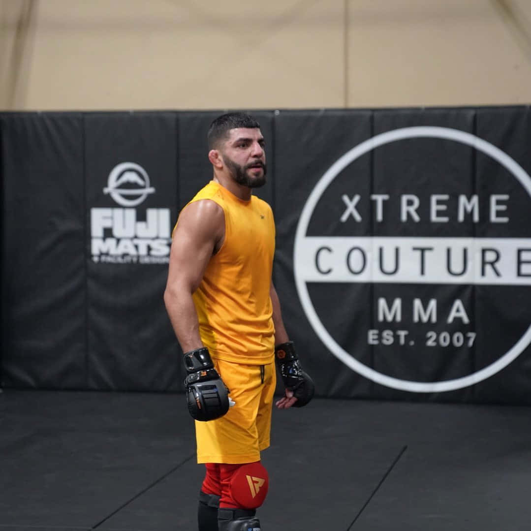 Fitness 桌布 壁紙 — 一位肌肉發達的MMA拳手身穿黃色裝備，專注地站在黑色墊子上，身後有'XTREME COUTURE MMA'品牌標誌。