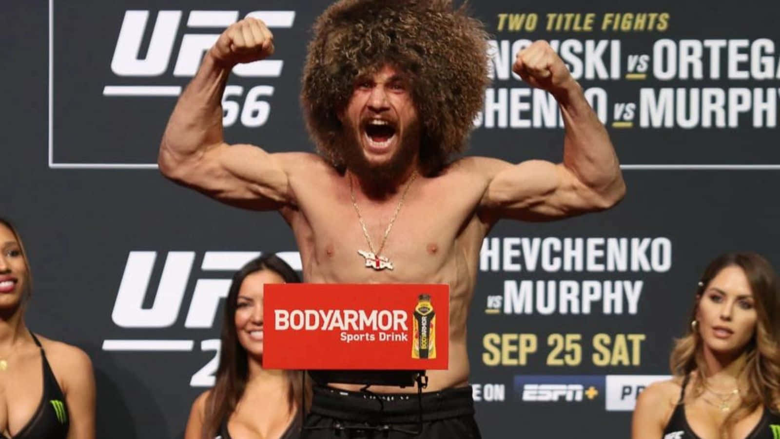 桌布 壁紙 — 一位肌肉發達的UFC拳手，擁有狂野的捲髮，在稱重活動中勝利地彎曲雙臂，附近有宣傳橫幅和擂台女郎。