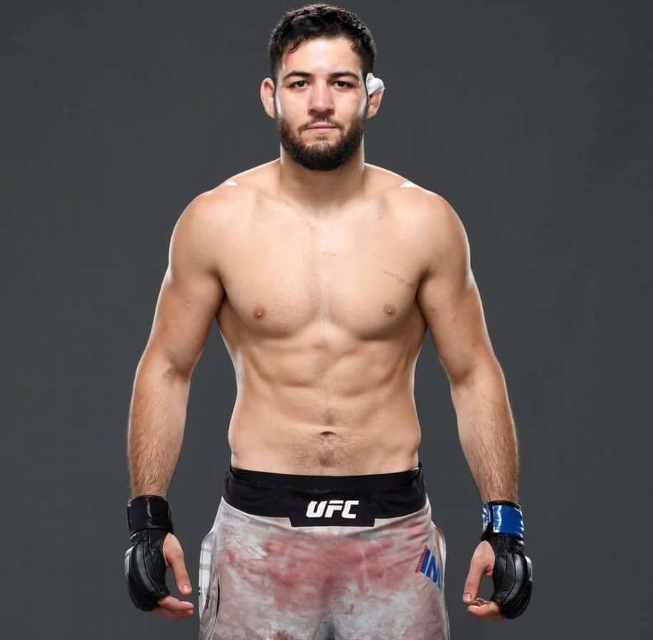運動 桌布 壁紙 — 一位肌肉發達的UFC拳手，留著鬍鬚，穿著沾有血跡的短褲，自信地站在深灰色工作室背景前。