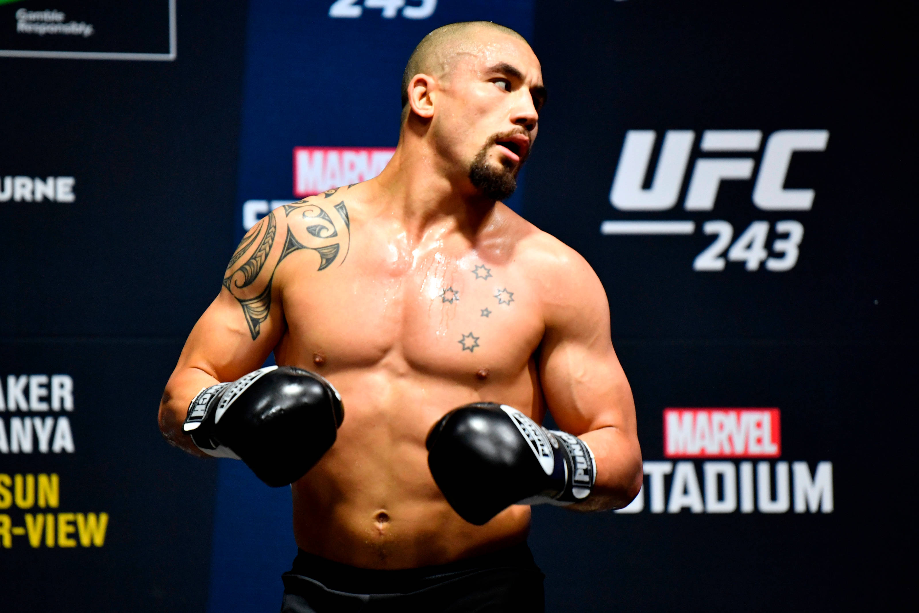 UFC 桌布 壁紙 — 一位肌肉發達的UFC拳手，身上有刺青，戴著黑色手套，在藍色活動背景前充滿氣勢地站立。