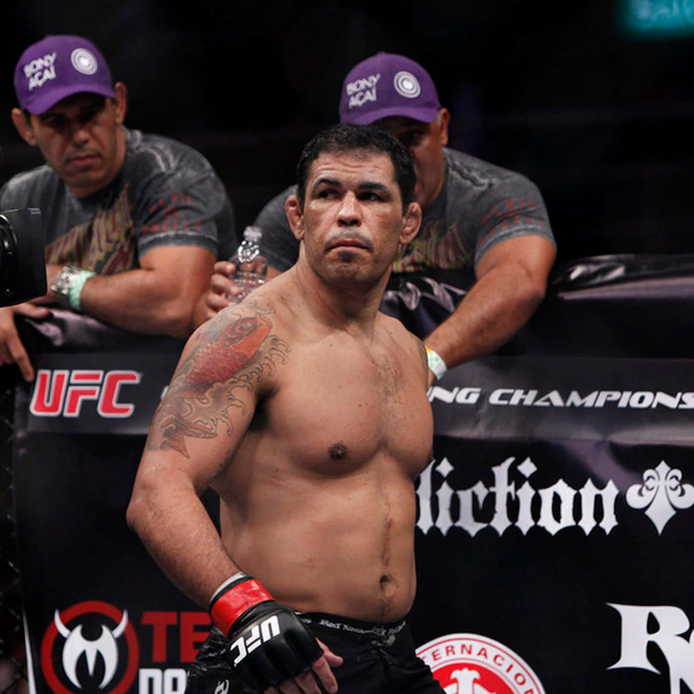 MMA 桌布 壁紙 — 一位肌肉發達的UFC拳手，身上有紅色紋身，在八角籠內凝視鏡頭，身後有戴紫色帽子的教練。