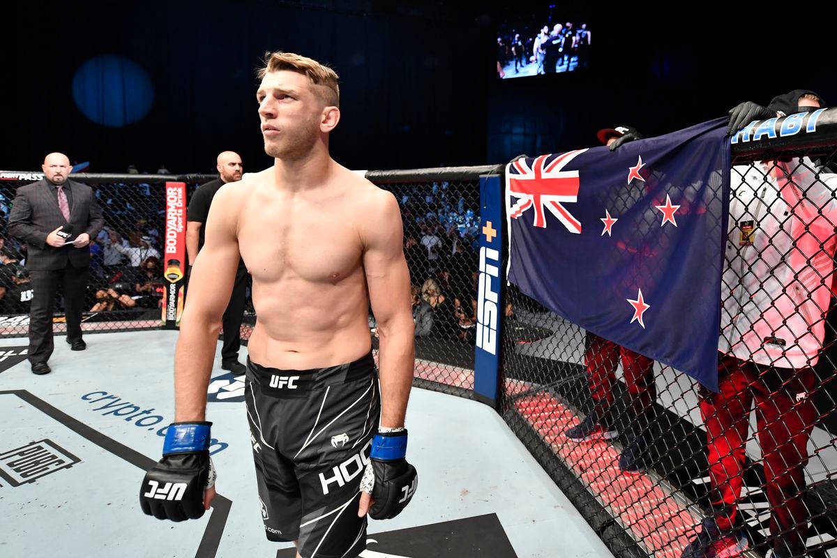 UFC 桌布 壁紙 — 一位肌肉發達的UFC拳手在八角籠內赤裸上身，身後圍欄上掛著紐西蘭國旗。