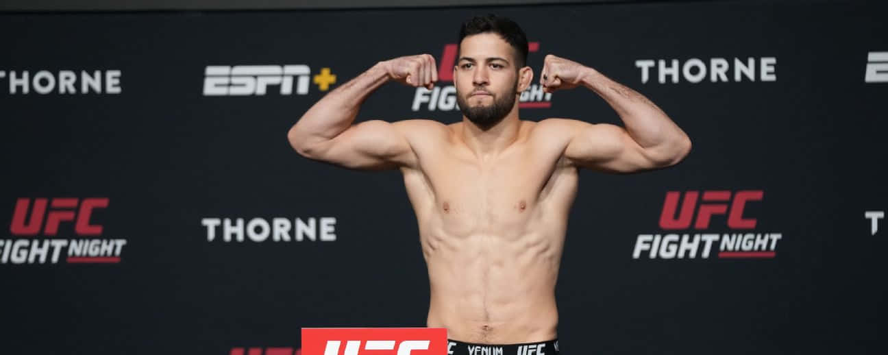 MMA 桌布 壁紙 — 一位肌肉發達的UFC拳手在品牌黑色背景前，自信地彎曲手臂進行稱重活動。