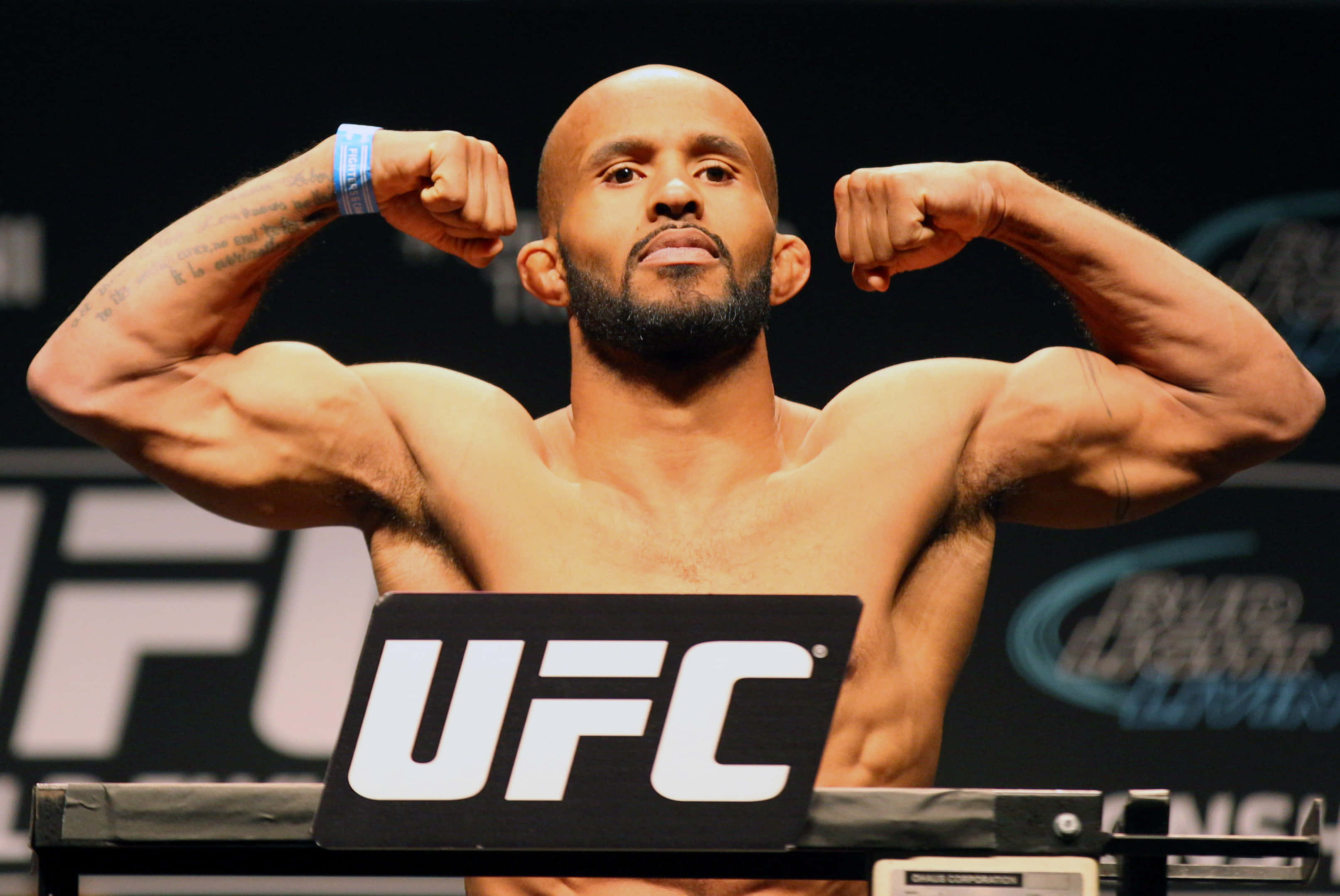 UFC 桌布 壁紙 — 一位肌肉發達的UFC拳手在明亮的場館燈光下，於稱重活動中集中精神彎曲二頭肌。