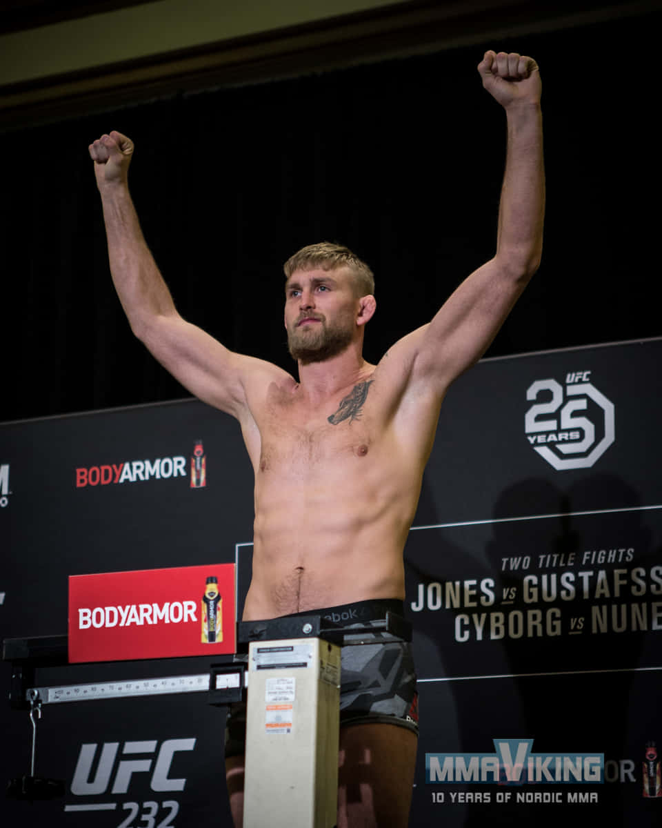 UFC 桌布 壁紙 — 一位肌肉發達的UFC拳手在明亮的場館燈光下，高舉拳頭勝利地站在體重計上。