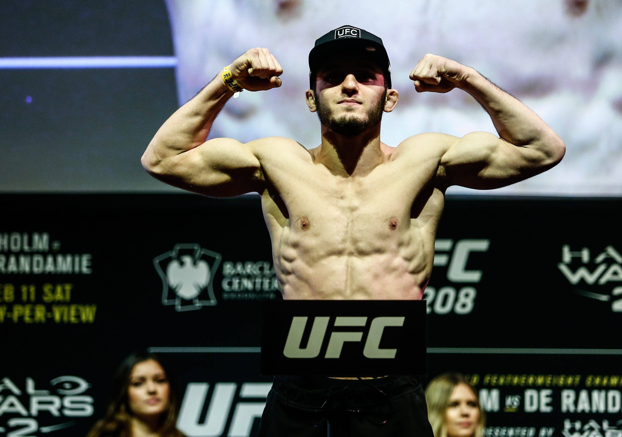UFC 桌布 壁紙 — 一位肌肉發達的UFC拳手在明亮的場館燈光下，於體重磅上彎曲手臂，身後有活動品牌標誌。