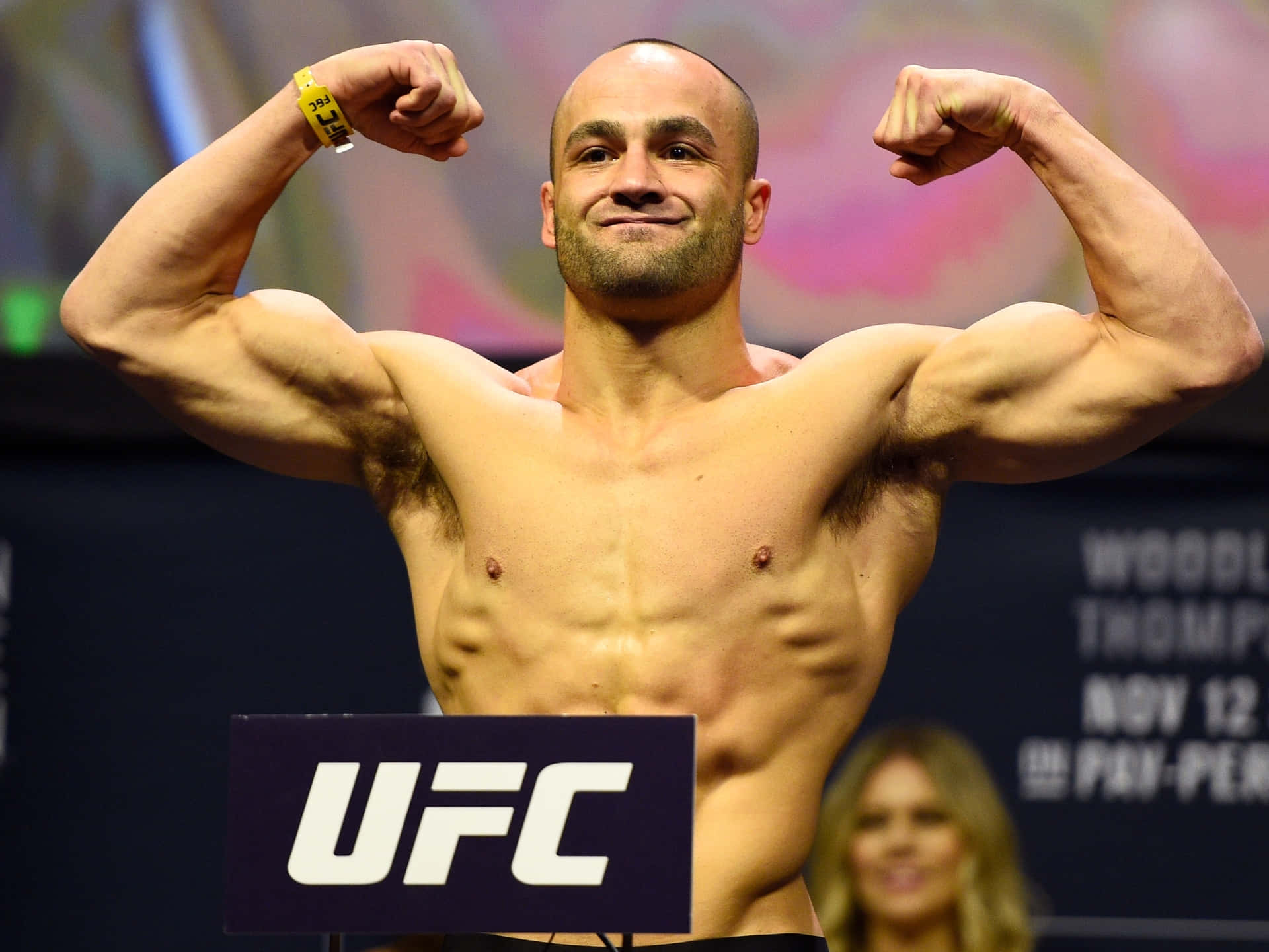 UFC 桌布 壁紙 — 一位肌肉發達的UFC拳手在明亮的場館燈光下，於稱重活動中自信地微笑並彎曲手臂。