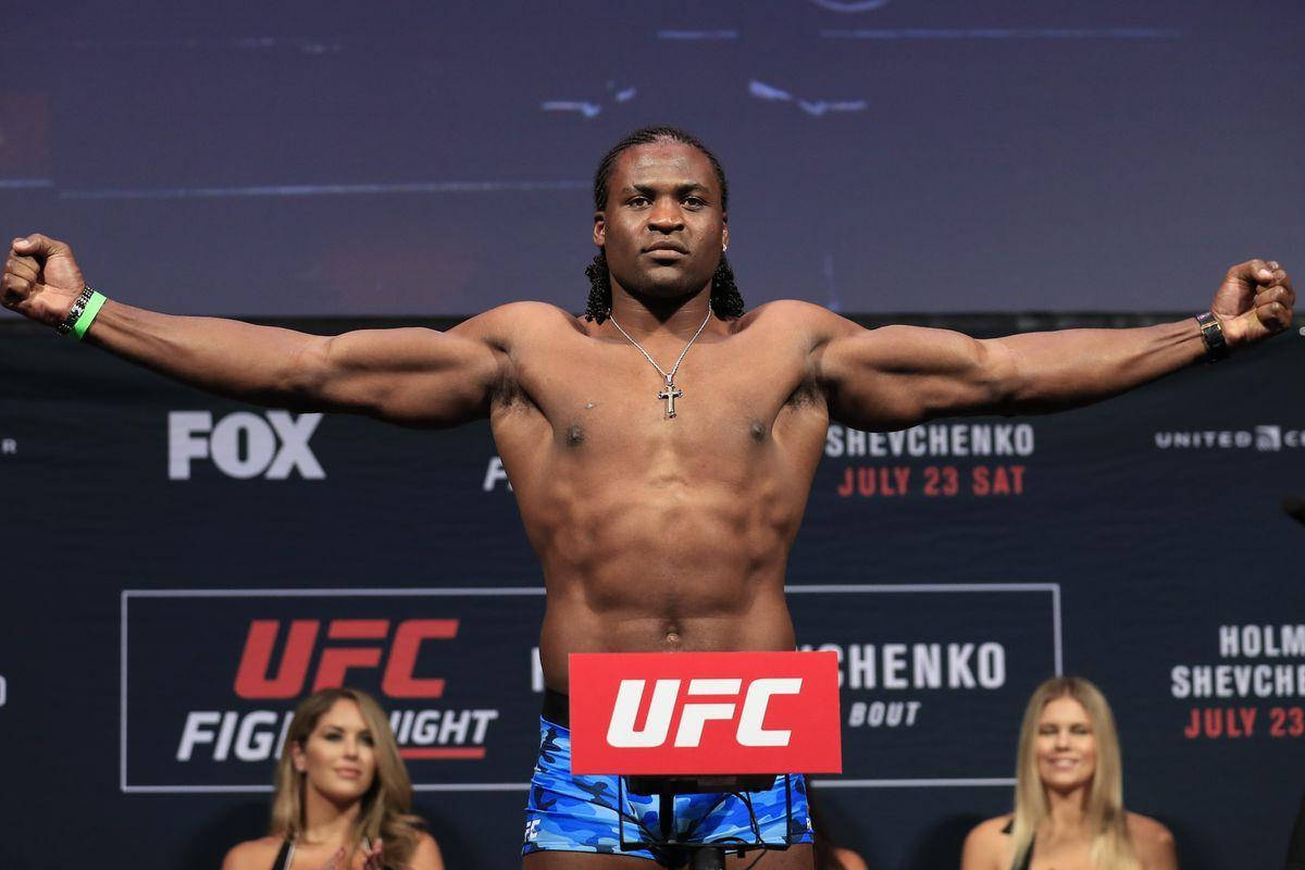 MMA 桌布 壁紙 — 一位肌肉發達的UFC拳手在明亮的場館燈光下，自信地張開雙臂站在體重磅上進行稱重活動。