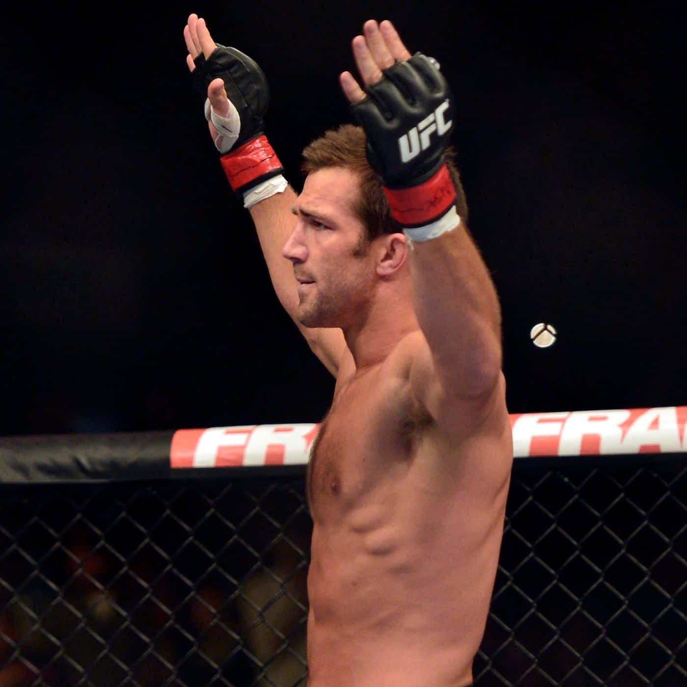 UFC 桌布 壁紙 — 一位肌肉發達的UFC拳手在昏暗的八角籠內舉起戴著手套的雙手慶祝勝利。