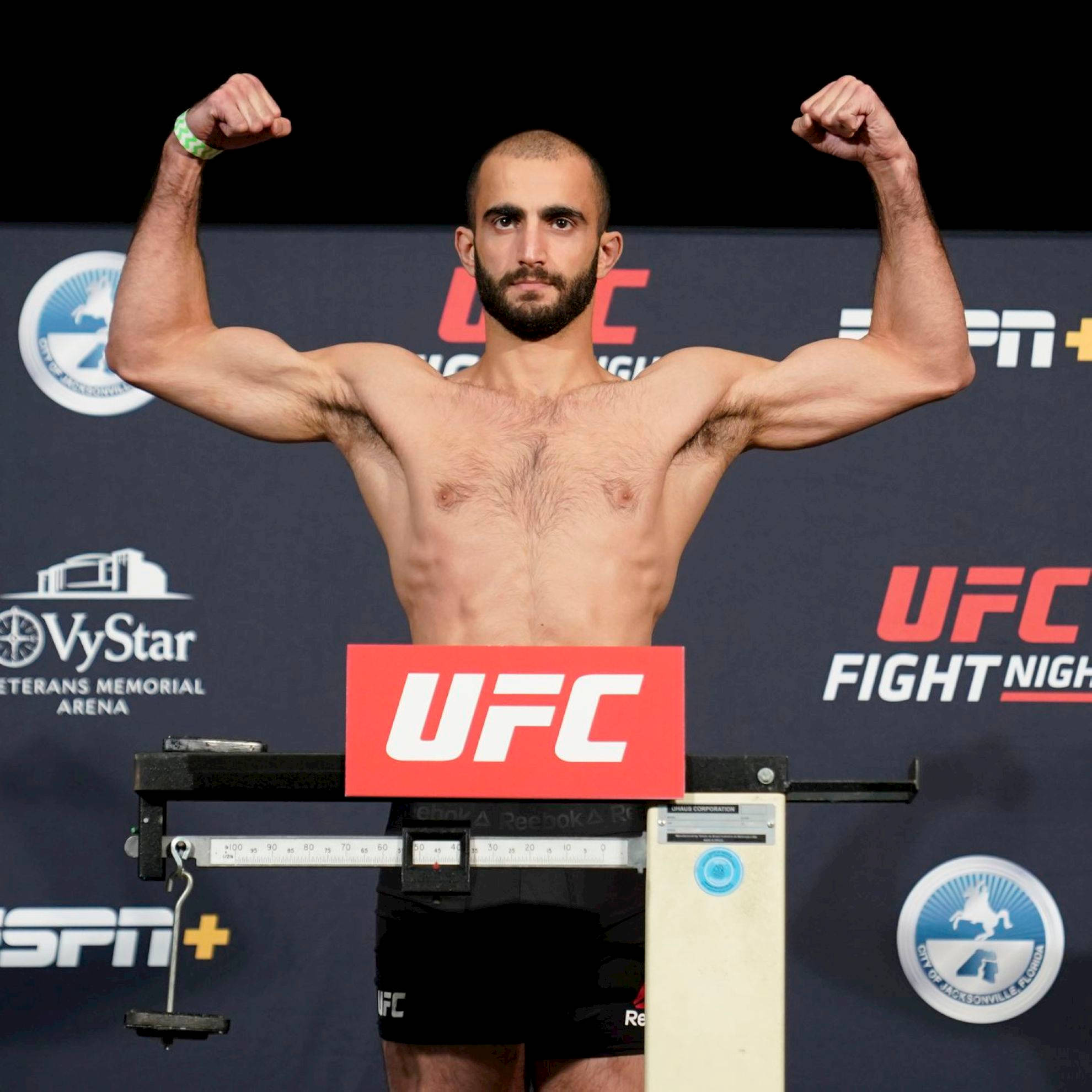 UFC 桌布 壁紙 — 一位肌肉發達的UFC拳手在有品牌背景的稱重活動中，在磅秤上彎曲手臂。