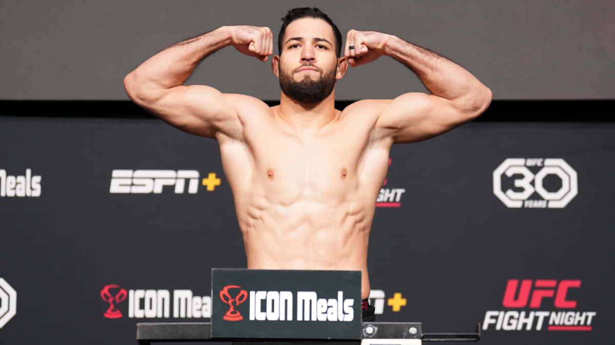 UFC 桌布 壁紙 — 一位肌肉發達的UFC拳手在秤重活動中，於體重磅上展示二頭肌，背後有ESPN和UFC品牌標誌。