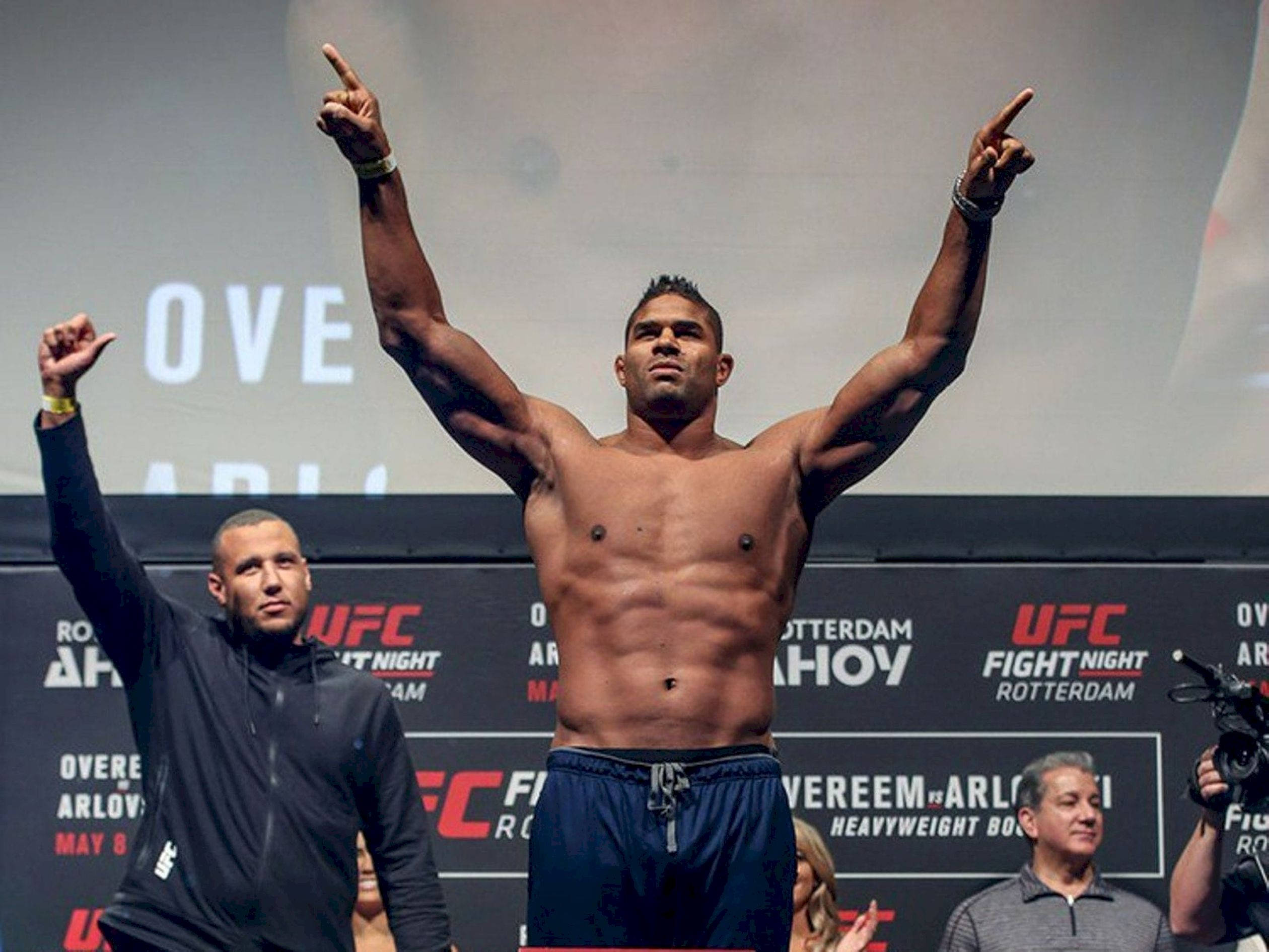 UFC 桌布 壁紙 — 一位肌肉發達的UFC拳手在稱重台上勝利地高舉雙臂，手指向上指。