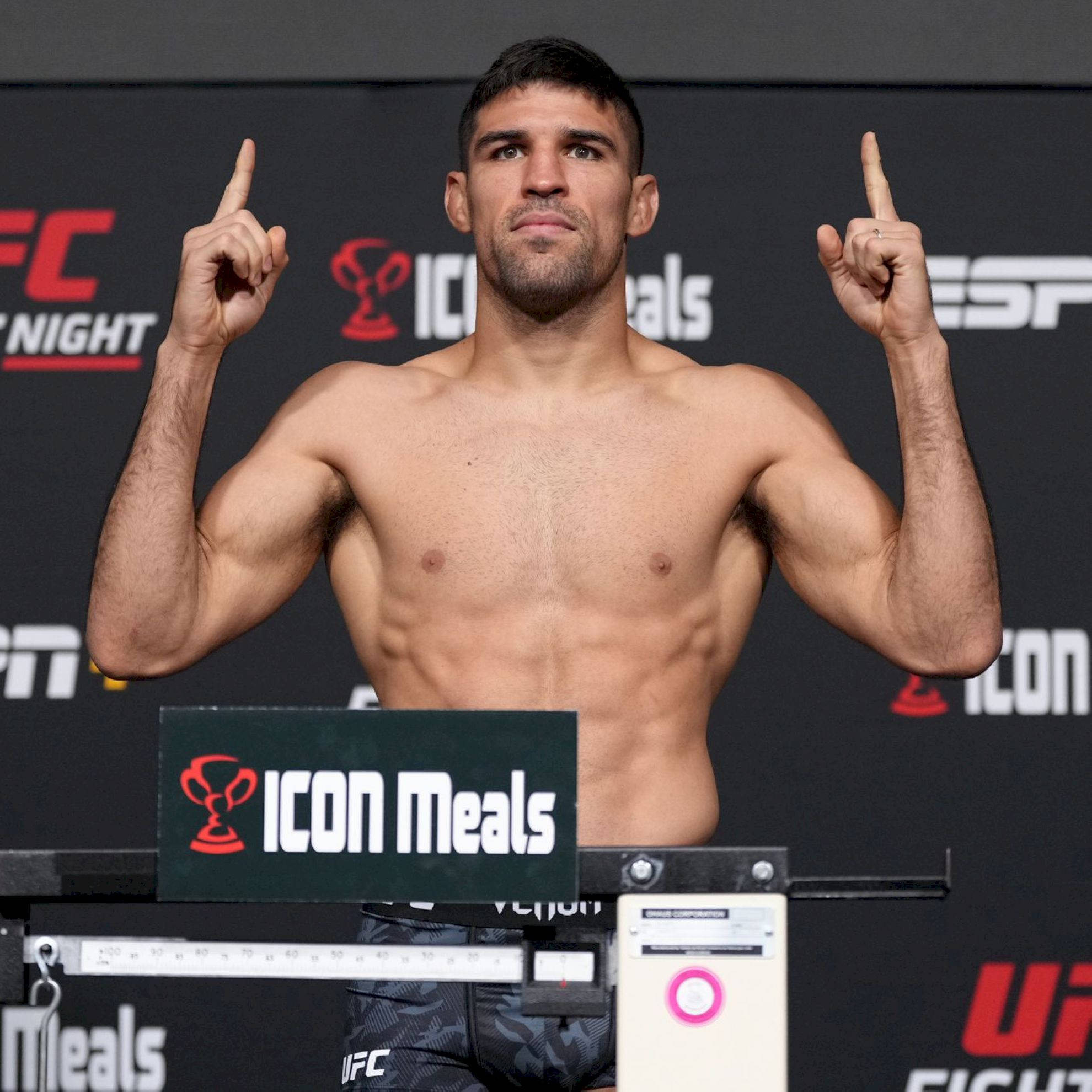 MMA 桌布 壁紙 — 一位肌肉發達的UFC拳手在稱重時赤裸上身站在體重磅上，雙食指向上指，神情專注。