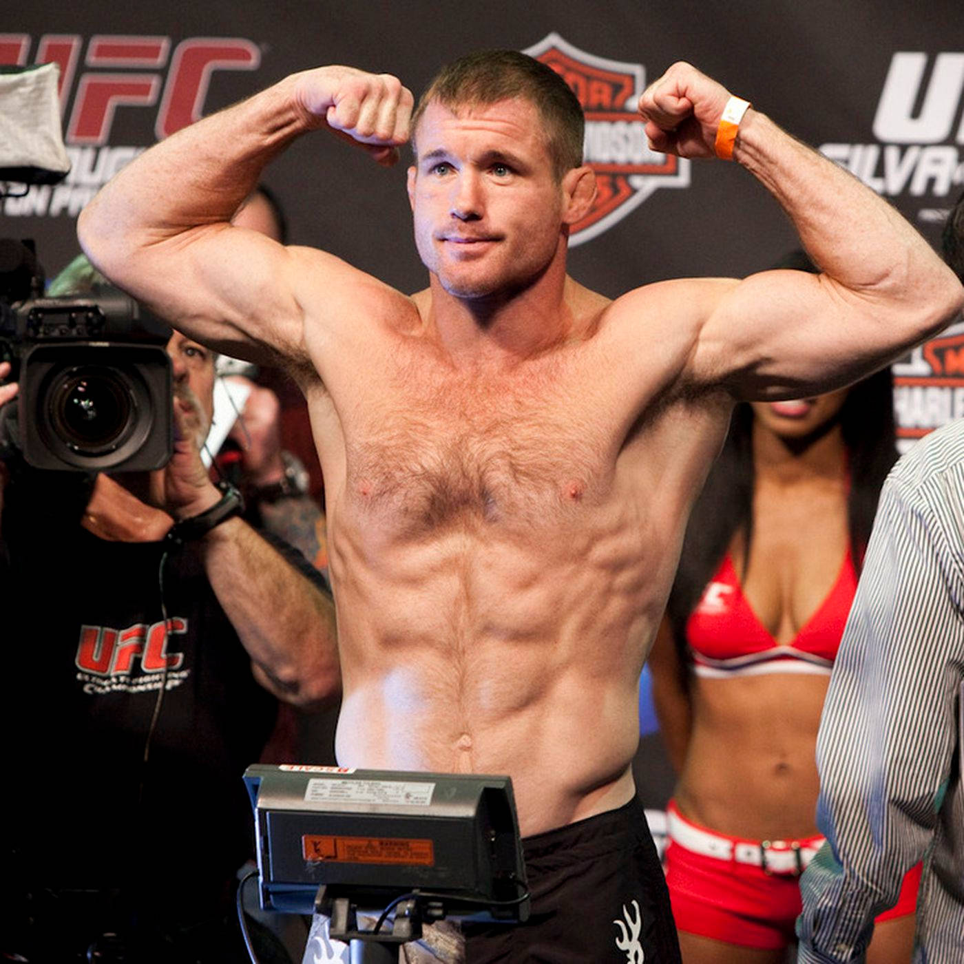 UFC 桌布 壁紙 — 一位肌肉發達的UFC拳手在稱重時彎曲手臂，背景有相機和啦啦隊。