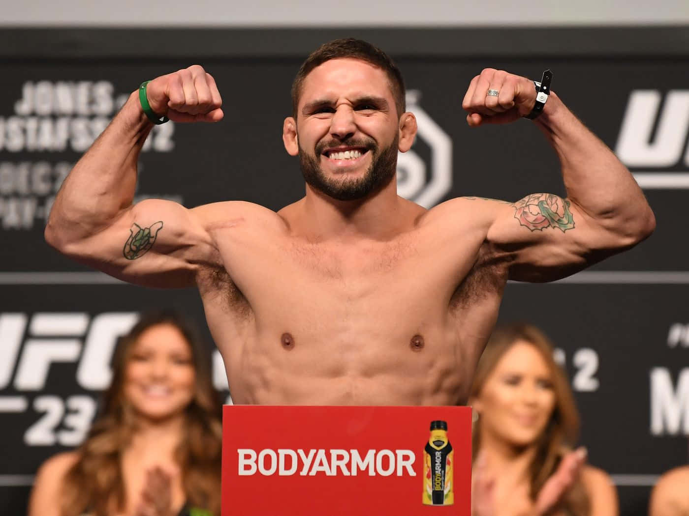 MMA 桌布 壁紙 — 一位肌肉發達的UFC拳手在稱重活動中彎曲手臂，咧嘴而笑，前面有一個紅色的BodyArmor標誌。