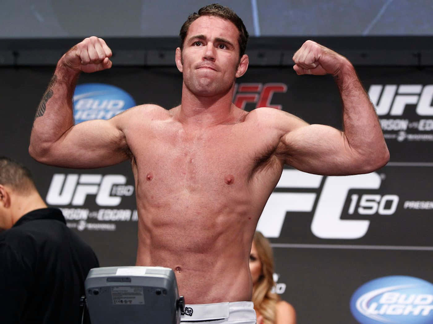 Athletic 桌布 壁紙 — 一位肌肉發達的UFC拳手在稱重活動中赤裸上身彎曲手臂，背景和體重秤帶有品牌標誌。