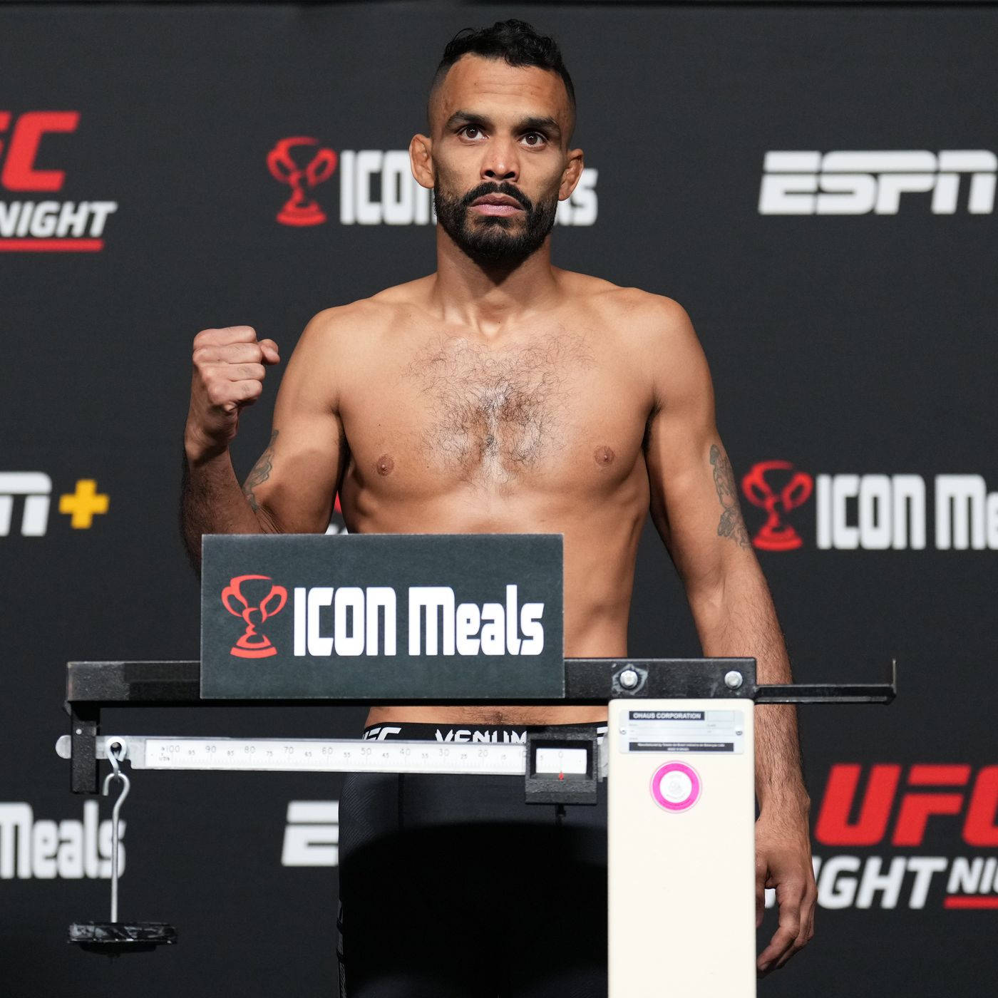 UFC 桌布 壁紙 — 一位肌肉發達的UFC拳手在稱重活動中赤裸上身站在體重磅上，身後有ESPN和ICON Meals的品牌標誌。