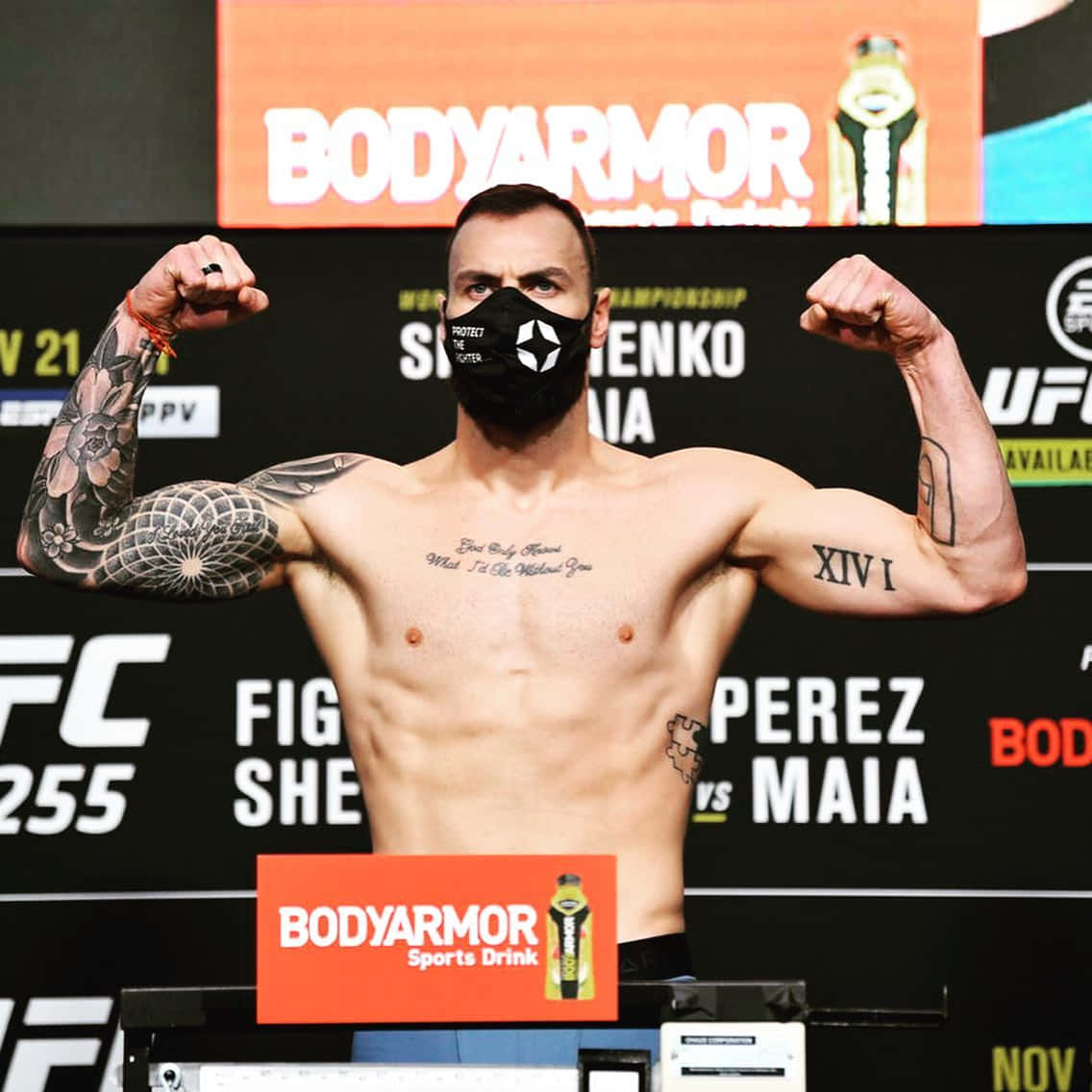 MMA 桌布 壁紙 — 一位肌肉發達的UFC拳手在稱重活動中彎曲手臂，戴著黑色口罩，身上有可見的刺青，身後有贊助商標誌。