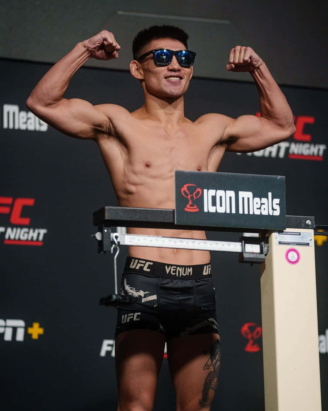 Athletic 桌布 壁紙 — 一位肌肉發達的UFC拳手在稱重活動中自信地站在體重計上展示肌肉，戴著太陽眼鏡和品牌短褲。