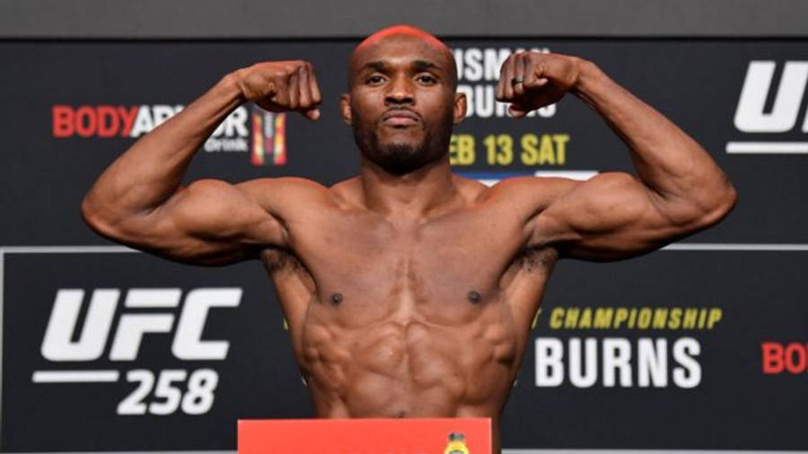 UFC 桌布 壁紙 — 一位肌肉發達的UFC拳手在稱重活動中，在體重磅上屈起手臂，身後有冠軍品牌標誌。