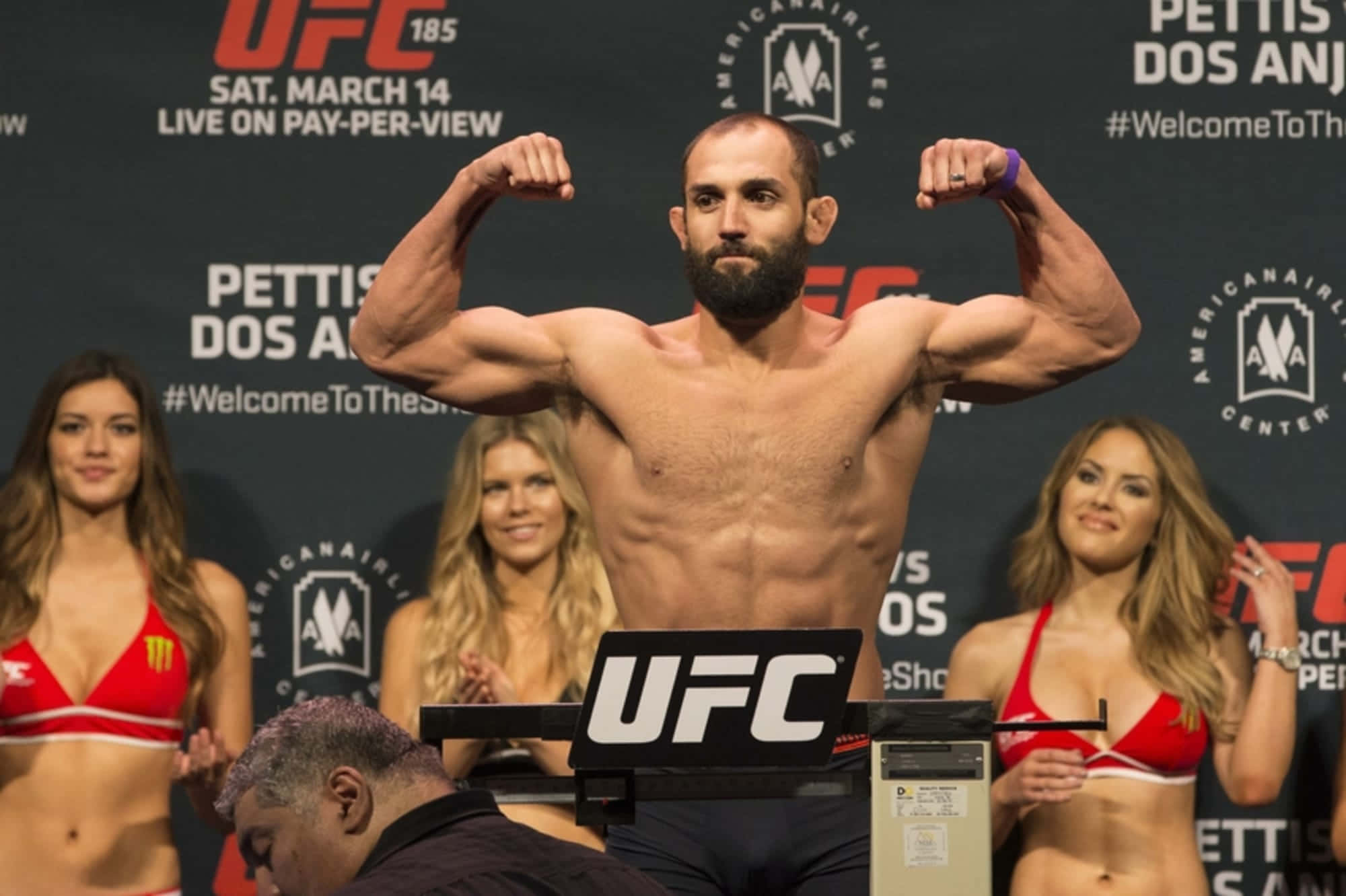 UFC 桌布 壁紙 — 一位肌肉發達的UFC拳手在稱重活動中於體重磅上屈起手臂，身後有三名身穿紅色比基尼、面帶微笑的擂台女郎。