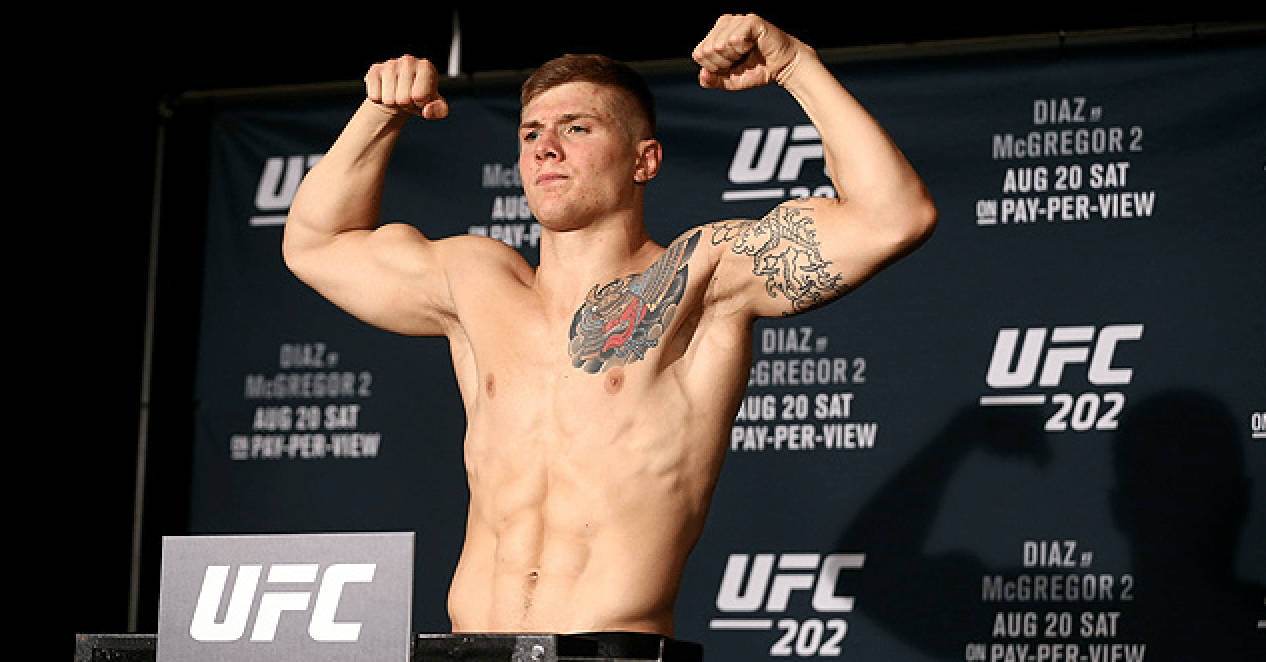 MMA 桌布 壁紙 — 一位肌肉發達的UFC拳手在稱重活動中，在磅秤上彎曲手臂，身後有宣傳橫幅。