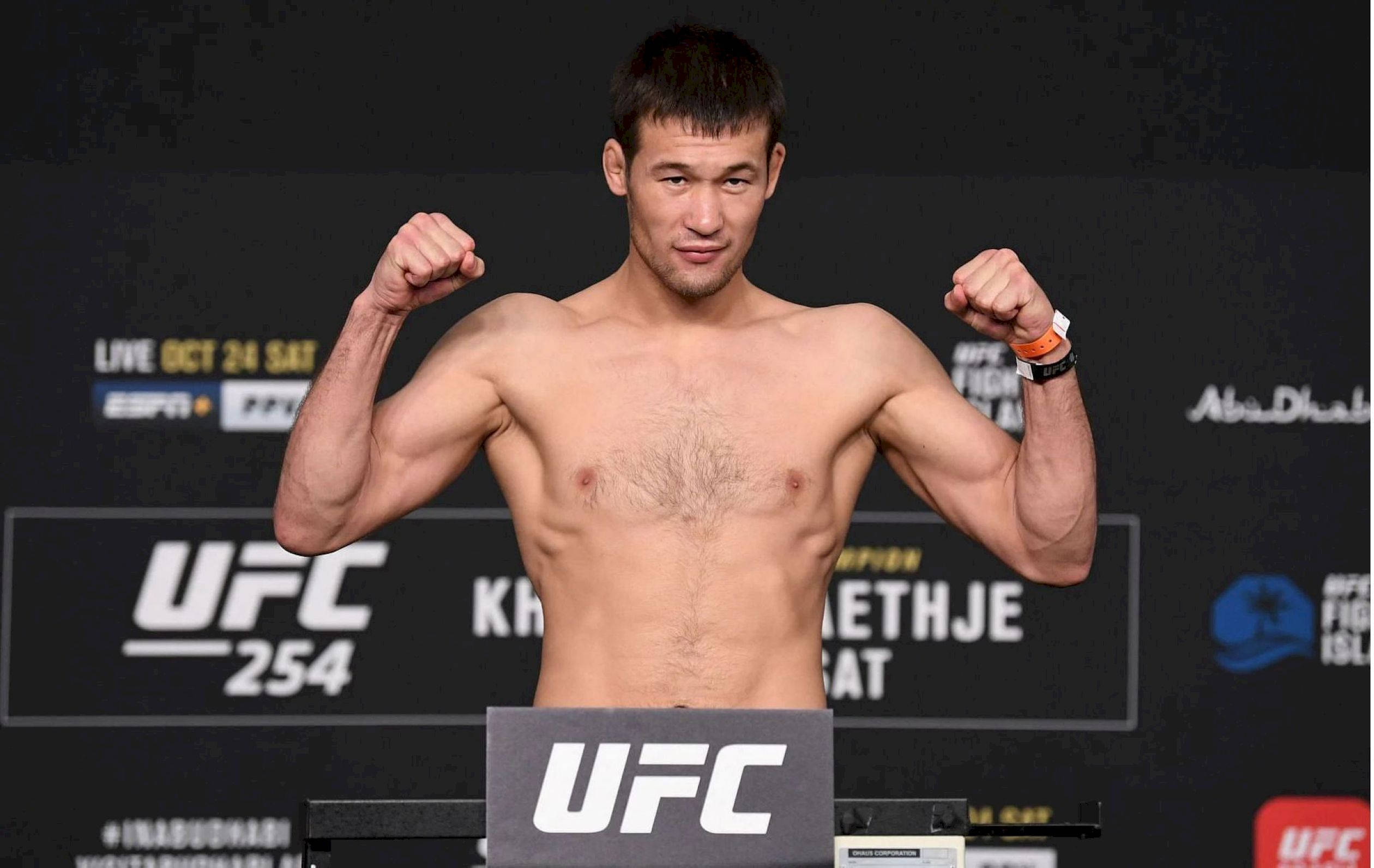 UFC 桌布 壁紙 — 一位肌肉發達的UFC拳手在稱重活動中彎曲雙臂，身後有冠軍旗幟。