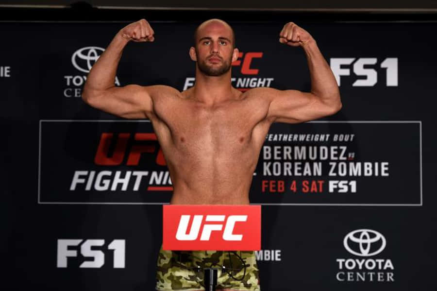 MMA 桌布 壁紙 — 一位肌肉發達的UFC拳手在稱重活動中赤裸上身彎曲手臂，背後可見標誌和拳手名字。