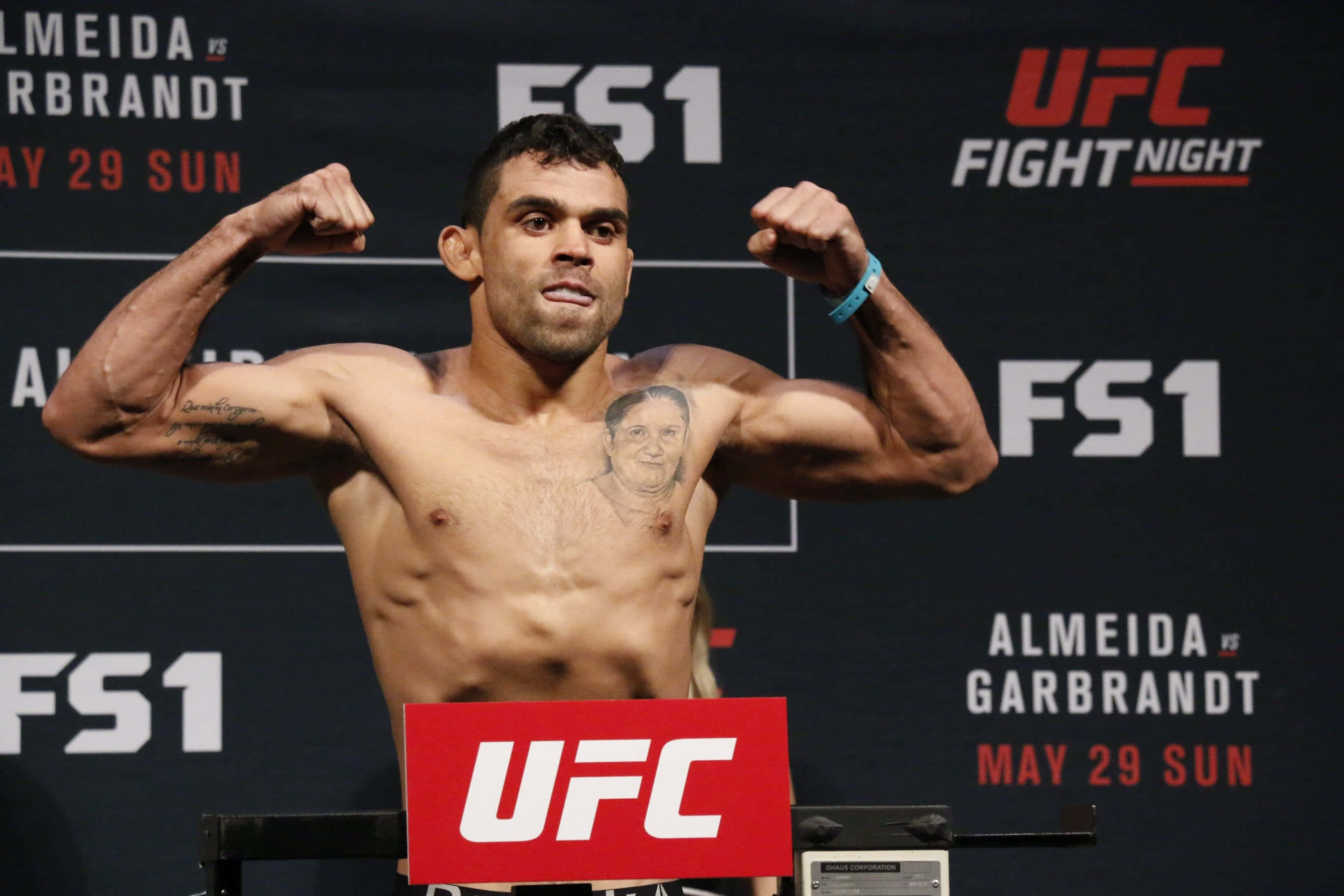 UFC 桌布 壁紙 — 一位肌肉發達的UFC拳手在稱重活動中彎曲手臂，身後有FS1和UFC Fight Night品牌標誌。