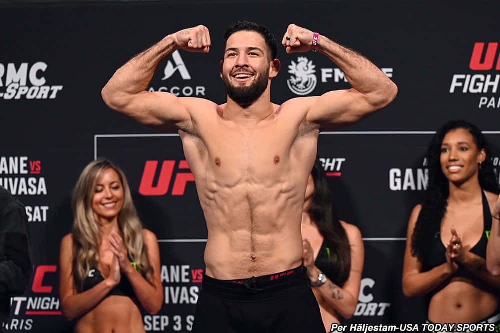 UFC 桌布 壁紙 — 一位肌肉發達的UFC拳手在稱重活動中自信地展示肌肉，身後有微笑的擂台女郎在鼓掌。