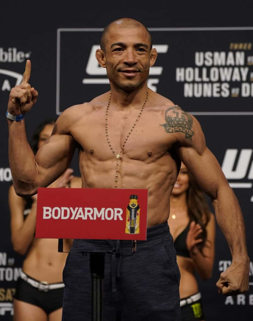 MMA 桌布 壁紙 — 一位肌肉發達的UFC拳手在稱重活動中站在體重磅上，微笑並向上指，旁邊有BodyArmor標誌。