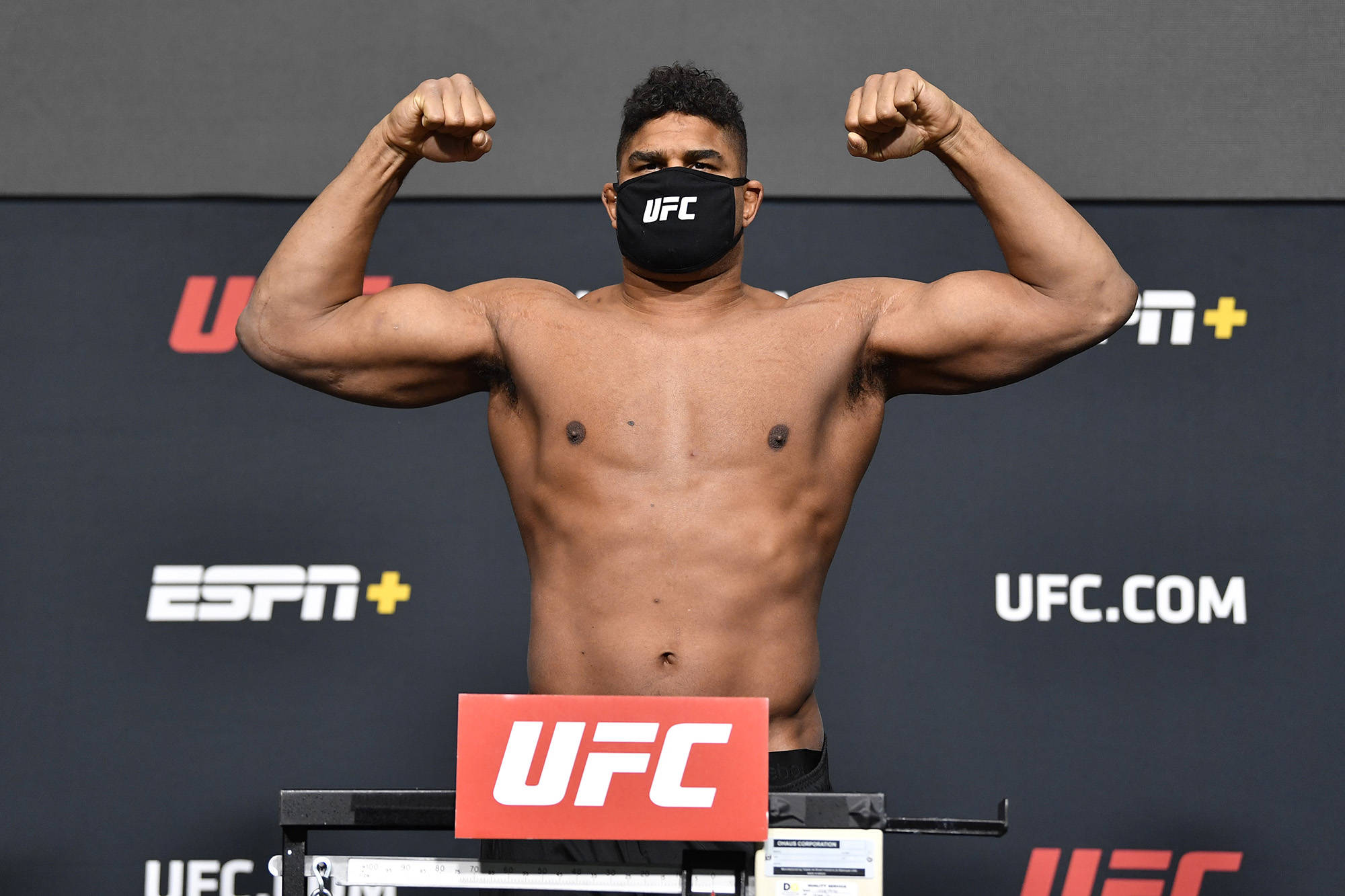 UFC 桌布 壁紙 — 一位肌肉發達的UFC拳手在稱重活動中戴著黑色口罩，彎曲手臂展示肌肉。