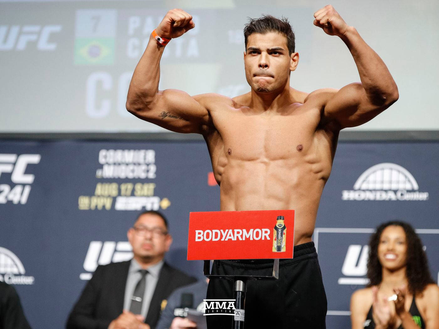 UFC 桌布 壁紙 — 一位肌肉發達的UFC拳手在稱重活動中，於有品牌橫幅和官員的背景前，在磅秤上彎曲手臂。