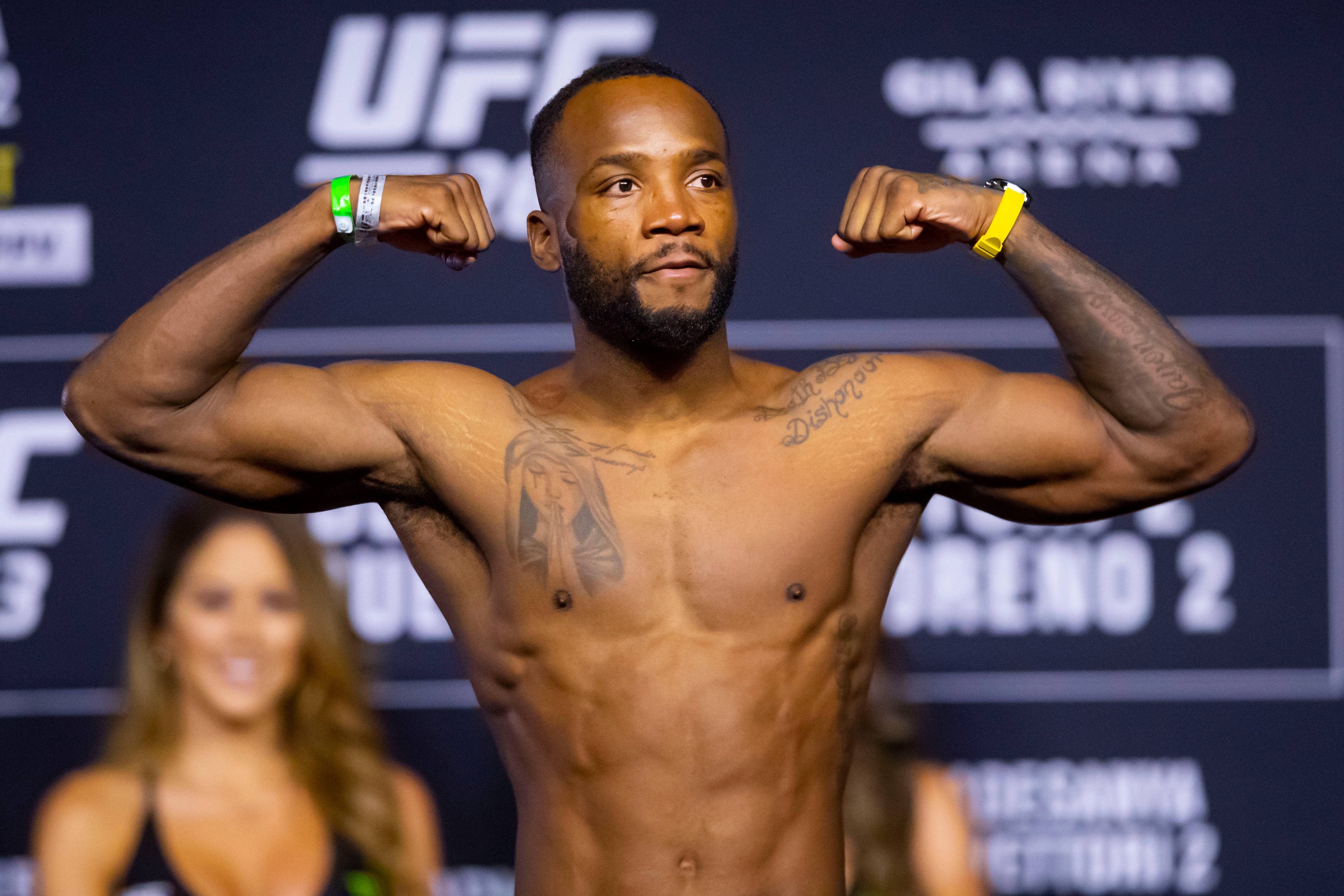 MMA 桌布 壁紙 — 一位肌肉發達的UFC拳手在稱重活動中彎曲手臂，背景有一位模糊的女性。