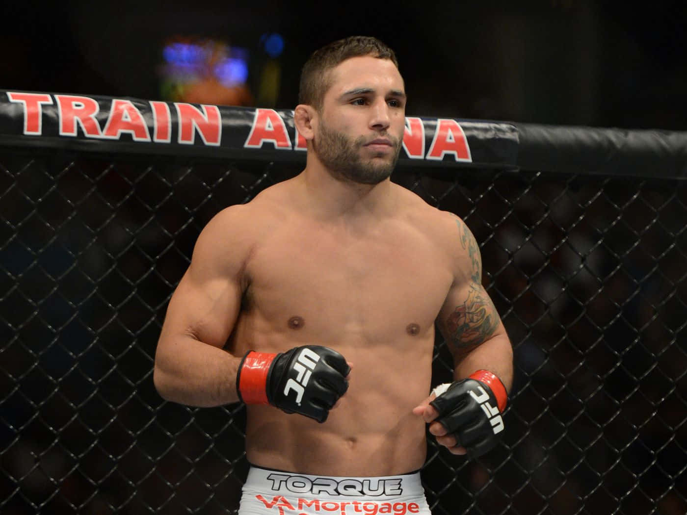 MMA 桌布 壁紙 — 一位肌肉發達的UFC拳手專注地站在籠內，身後圍欄上可見「TRAIN A」字樣。
