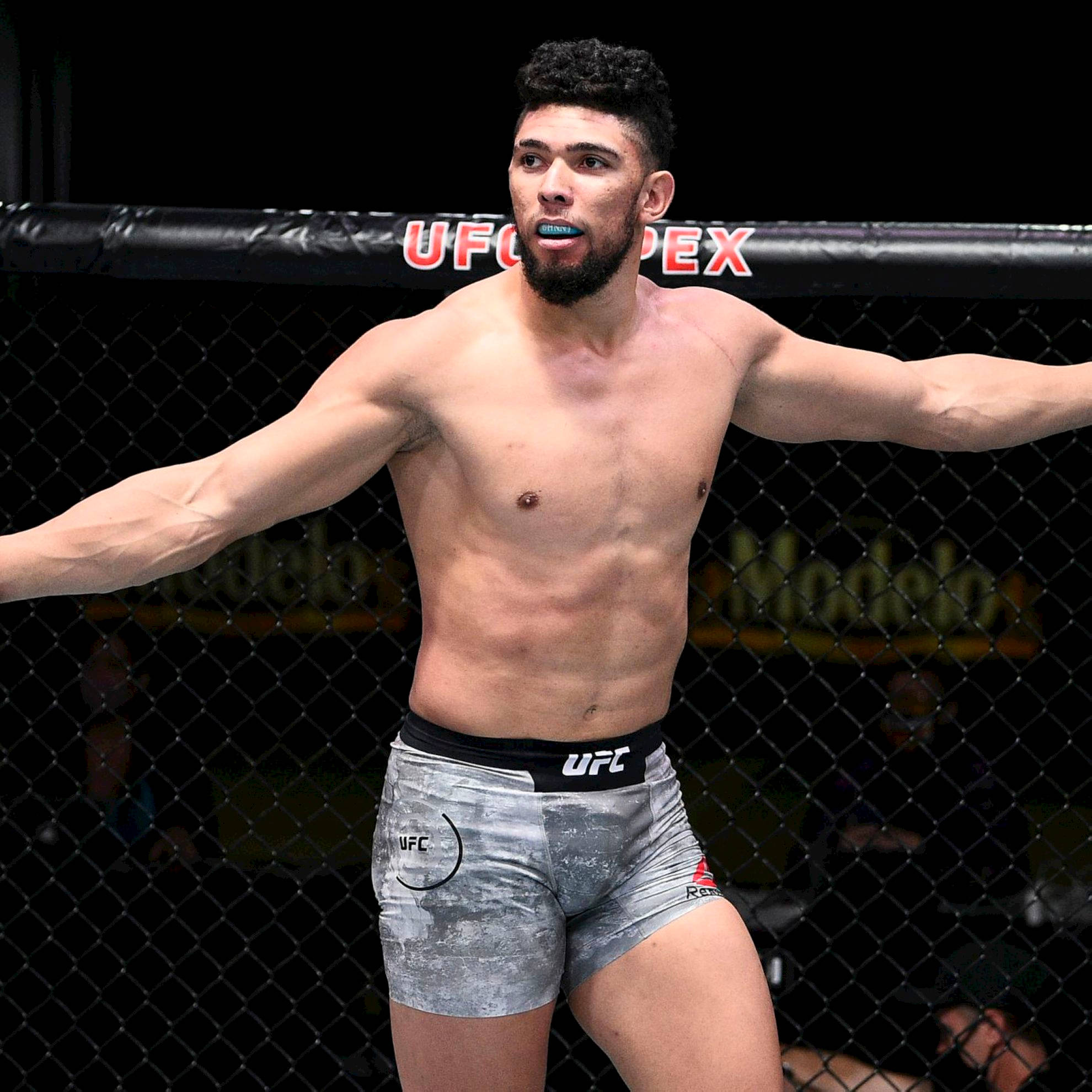 MMA 桌布 壁紙 — 一位肌肉發達的UFC拳手戴著藍色牙套，在八角籠中張開雙臂，勝利地站立。