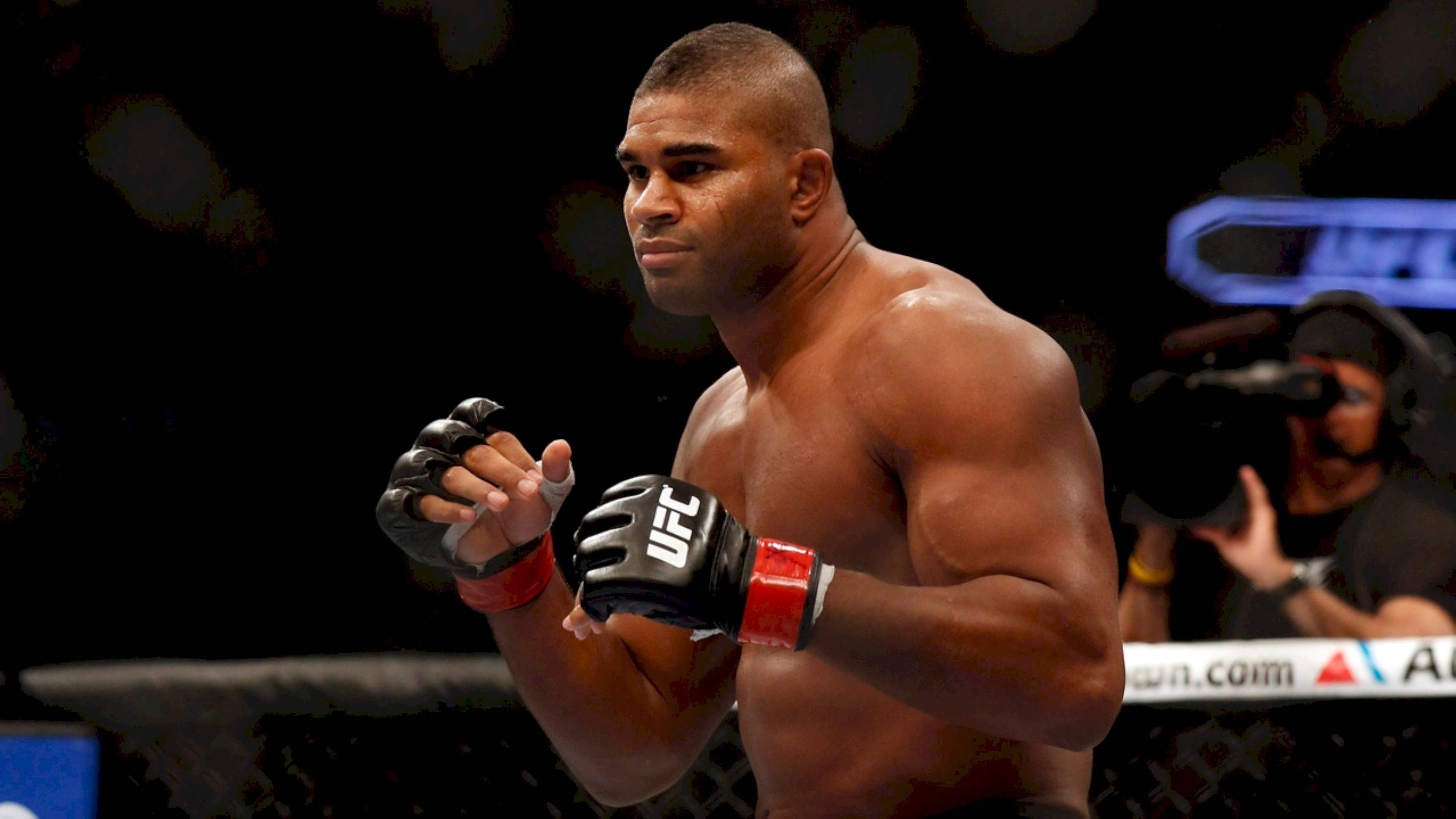 UFC 桌布 壁紙 — 一位肌肉發達的UFC拳手戴著黑色手套，在明亮的場館燈光下專注地站在八角籠中。