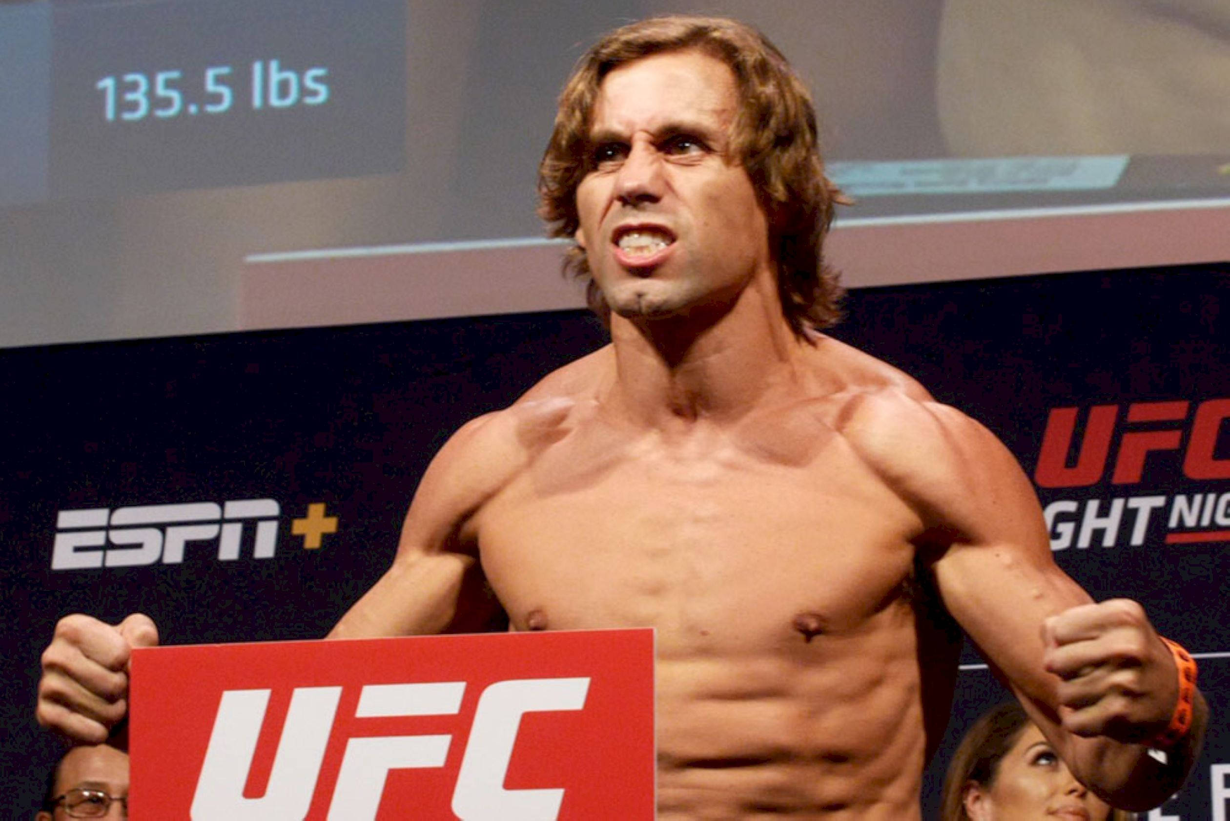MMA 桌布 壁紙 — 一位肌肉發達的UFC拳手留著長髮，在稱重活動中拿著紅色UFC標誌牌，表情猙獰。