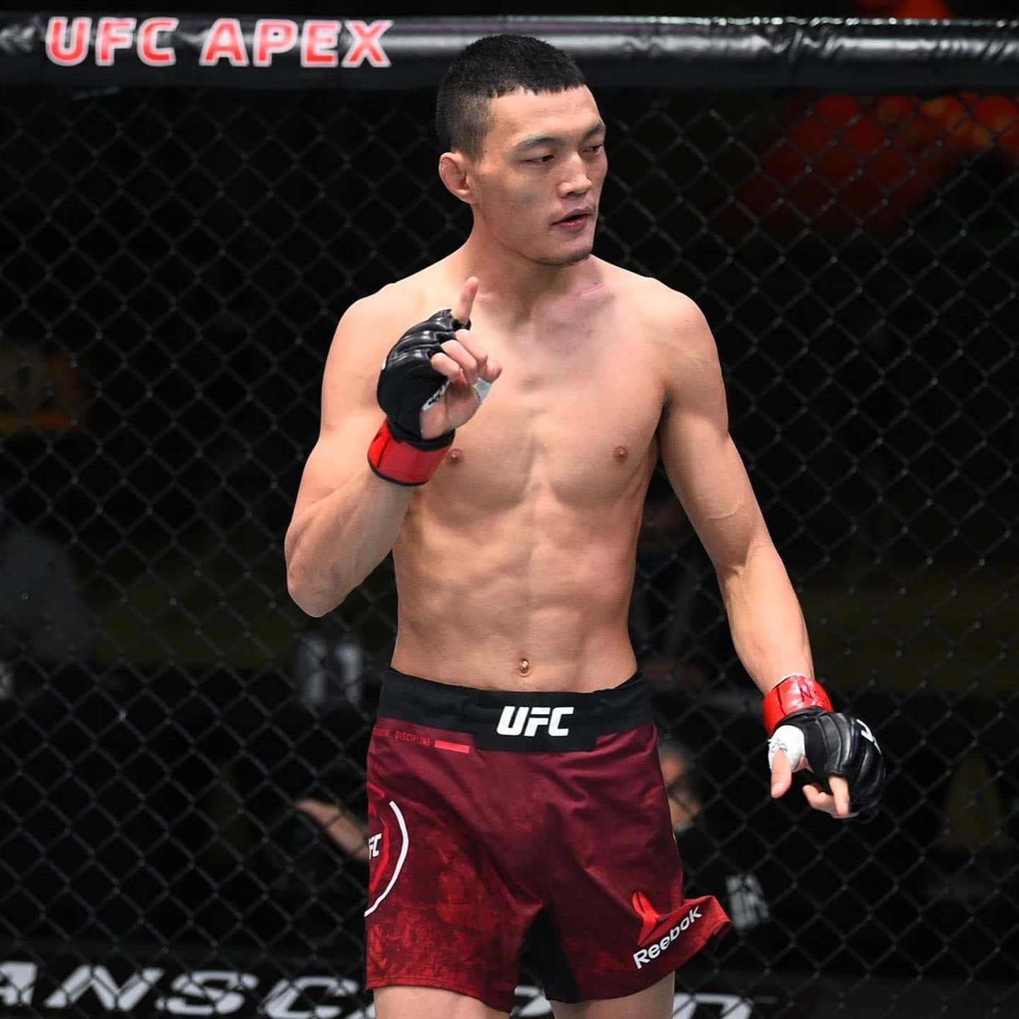 UFC 桌布 壁紙 — 一位肌肉發達的UFC拳手穿著紅色短褲，在明亮的場館燈光下，於UFC APEX的八角籠內自信地做手勢。