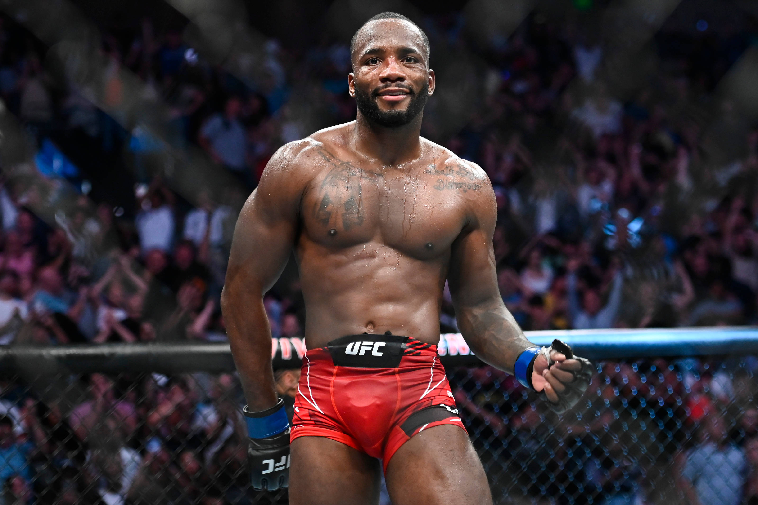 MMA 桌布 壁紙 — 一位肌肉發達的UFC拳手穿著紅色短褲，在八角籠中勝利站立，背後有歡呼的人群。
