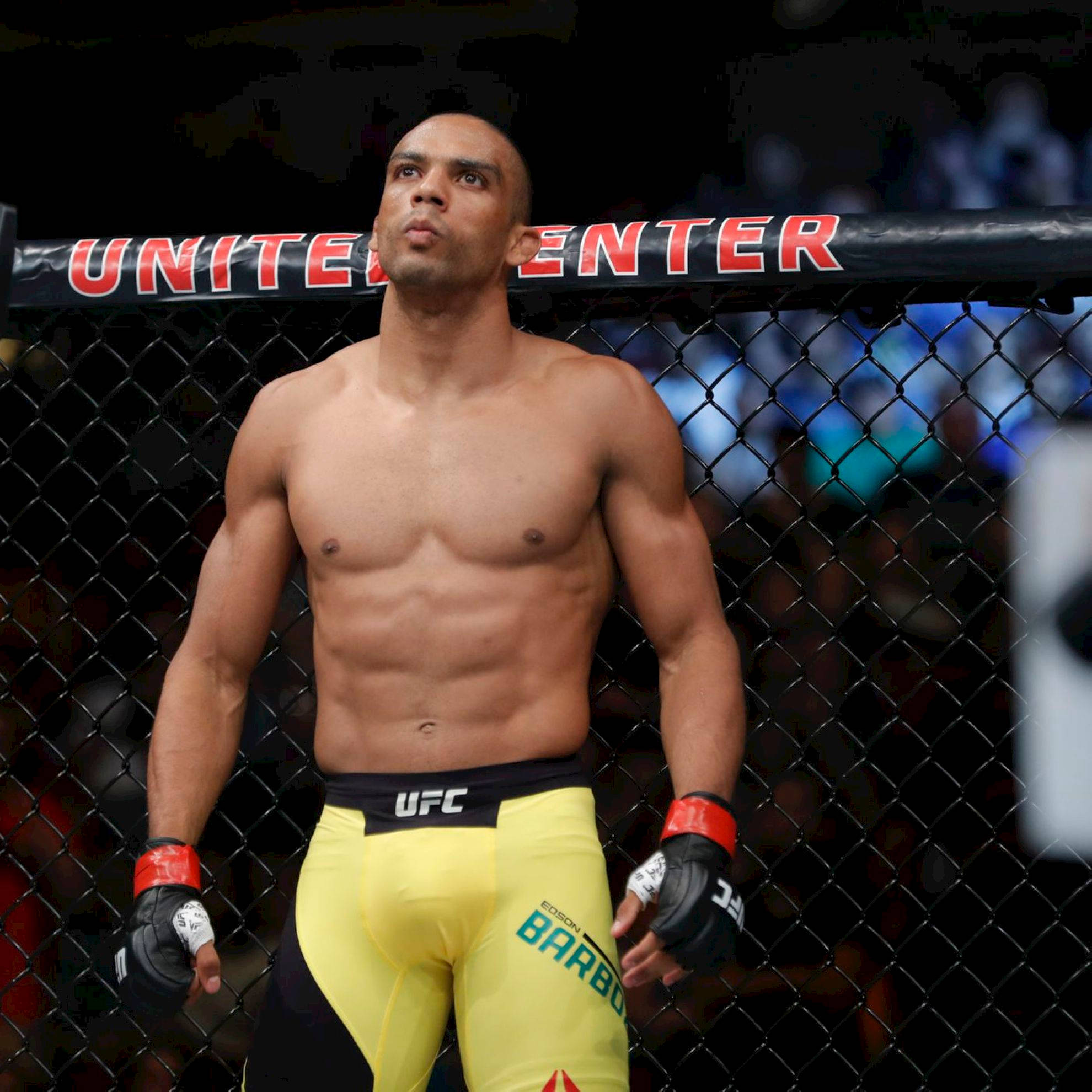UFC 桌布 壁紙 — 一位肌肉發達的UFC拳手穿著黃色短褲，站在八角籠內，專注地向上看。