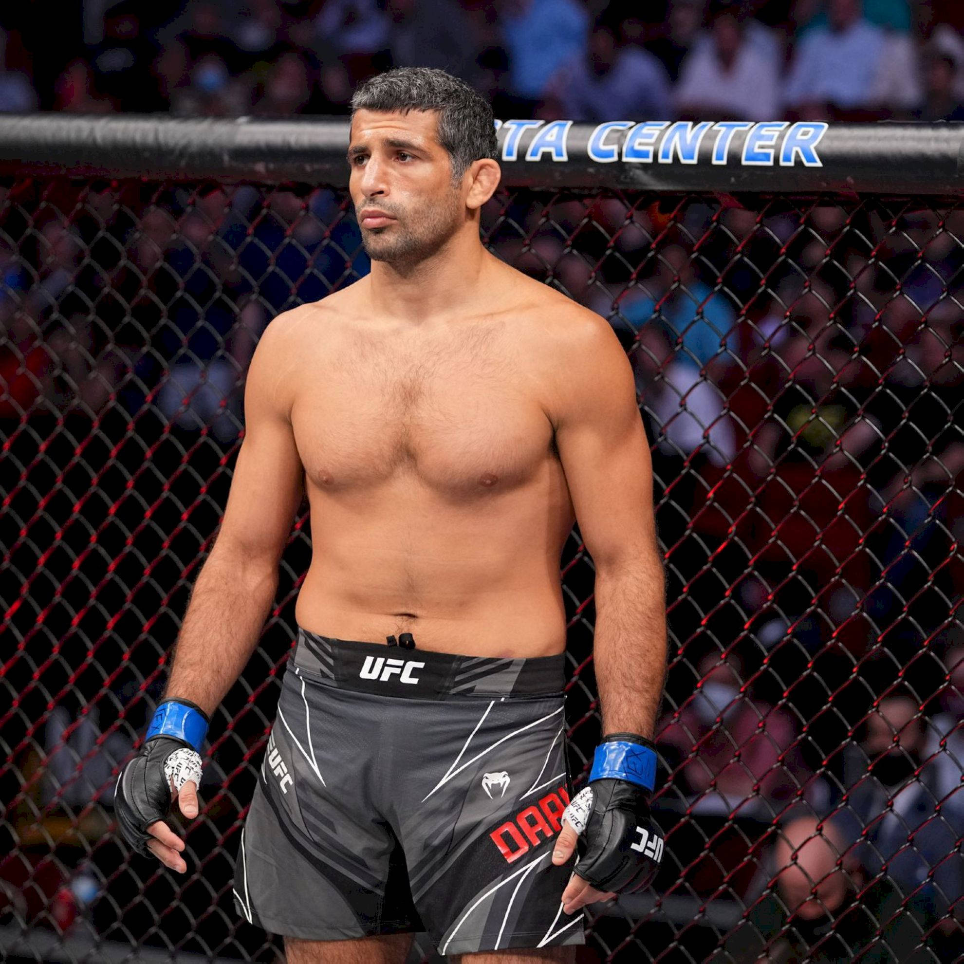 UFC 桌布 壁紙 — 一位肌肉發達的UFC拳手站在八角籠內，穿著黑色短褲和手套，神情專注。