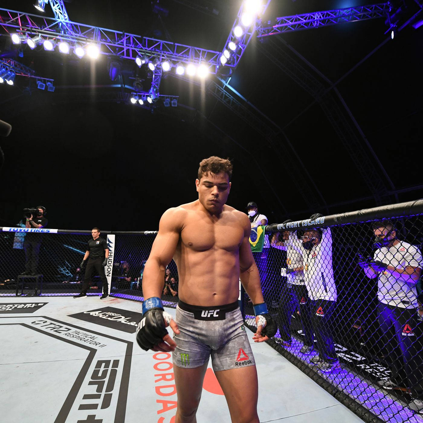 UFC 桌布 壁紙 — 一位肌肉發達的UFC拳手站在八角籠內，周圍有明亮的場館燈光、觀眾和官員。