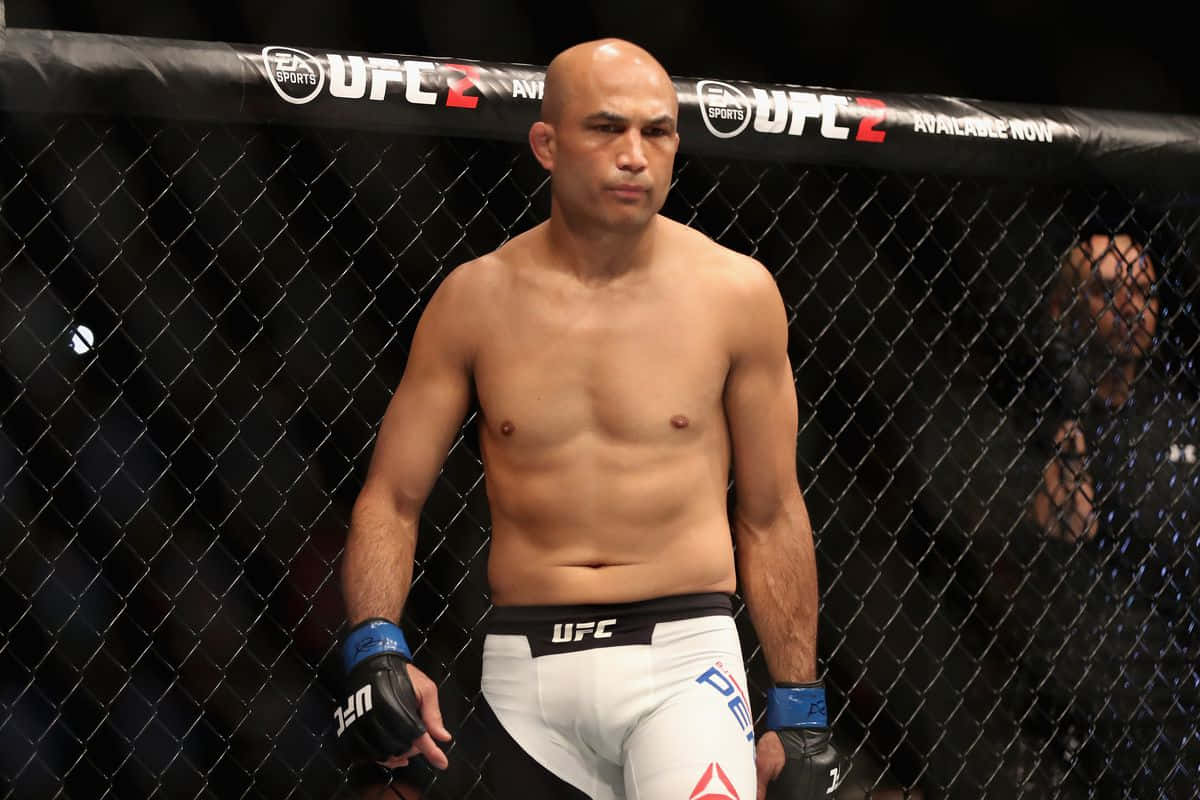 UFC 桌布 壁紙 — 一位肌肉發達的UFC拳手站在八角籠內，穿著白色短褲和藍色手套，神情專注。