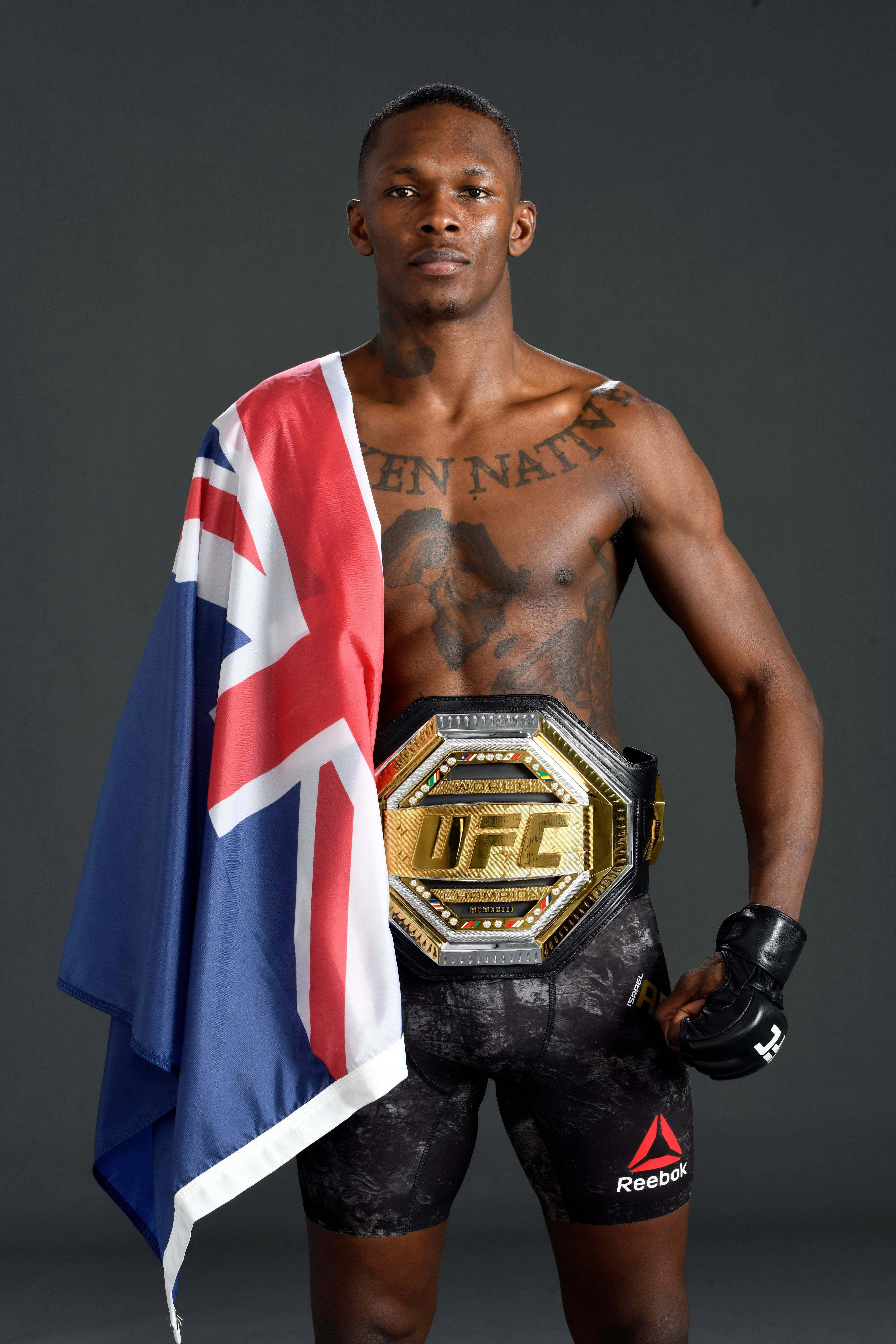 Adesanya 桌布 壁紙 — 一位肌肉發達的UFC拳手自信地站著，腰間繫著冠軍腰帶，披著英國國旗，背景是黑暗的攝影棚。